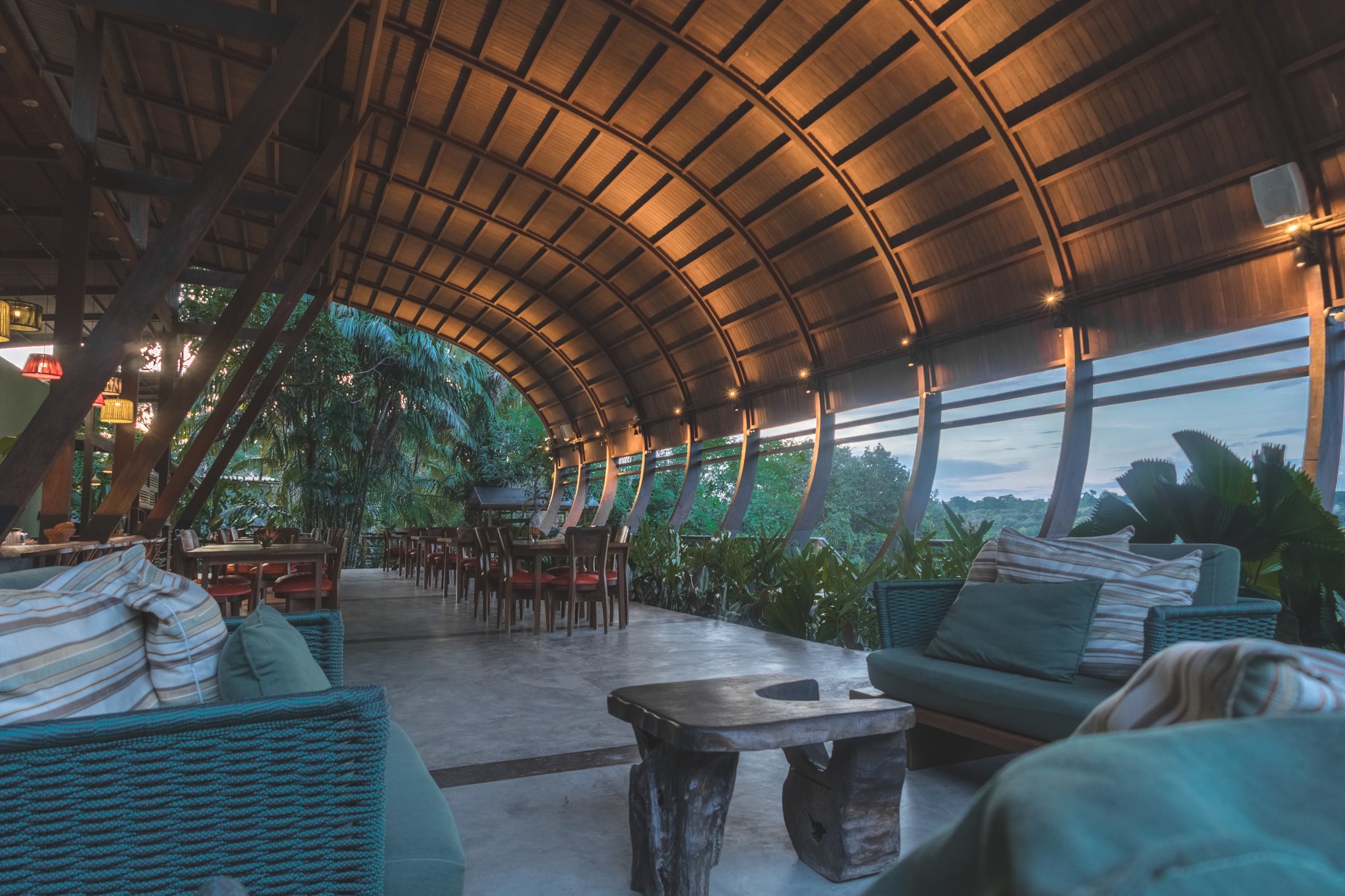 Dine under the jungle canopy — where bold architecture meets the Amazon at dusk. Curved wooden barrel-vault ceiling arching over the open-air Mirante do Gavião restaurant lounge, with rattan sofas, rustic log coffee table, dining tables, and a panoramic wall of tropical rainforest at dusk