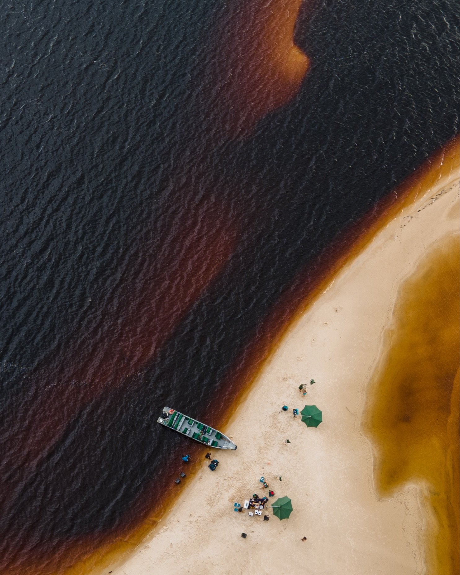 Where dark Rio Negro waters meet white Amazonian sands — a fleeting paradise near Mirante do Gavião. Aerial drone view of a small motorboat moored on a pale sandy river beach at the edge of the dark tannin-stained Rio Negro, with green umbrellas and guests gathered at the Mirante do Gavião Amazon lodge river stop