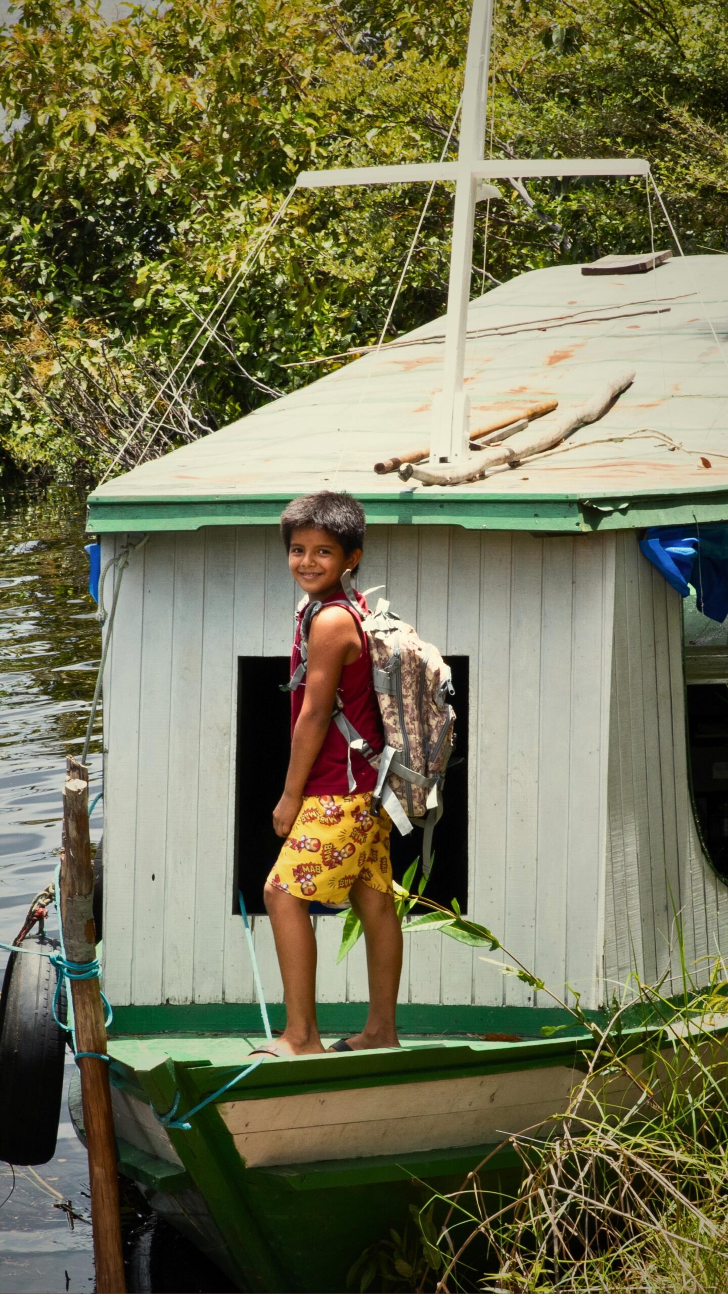 River life up close — a local child ready for the day along the Amazon shore. A smiling boy wearing a backpack stands on the deck of a small wooden houseboat moored along a lush Amazon riverbank, near the Mirante do Gavião riverside communities