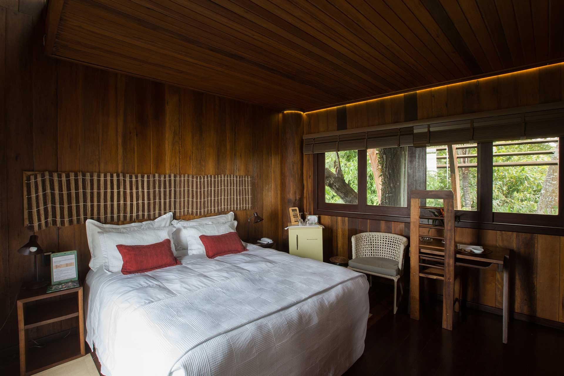 Wake up surrounded by the Amazon — rich hardwood walls and jungle views straight from your bed. Warm wooden-panelled bedroom at Mirante do Gavião with a king-size bed dressed in white linen, red accent cushions, and large windows overlooking dense Amazon rainforest