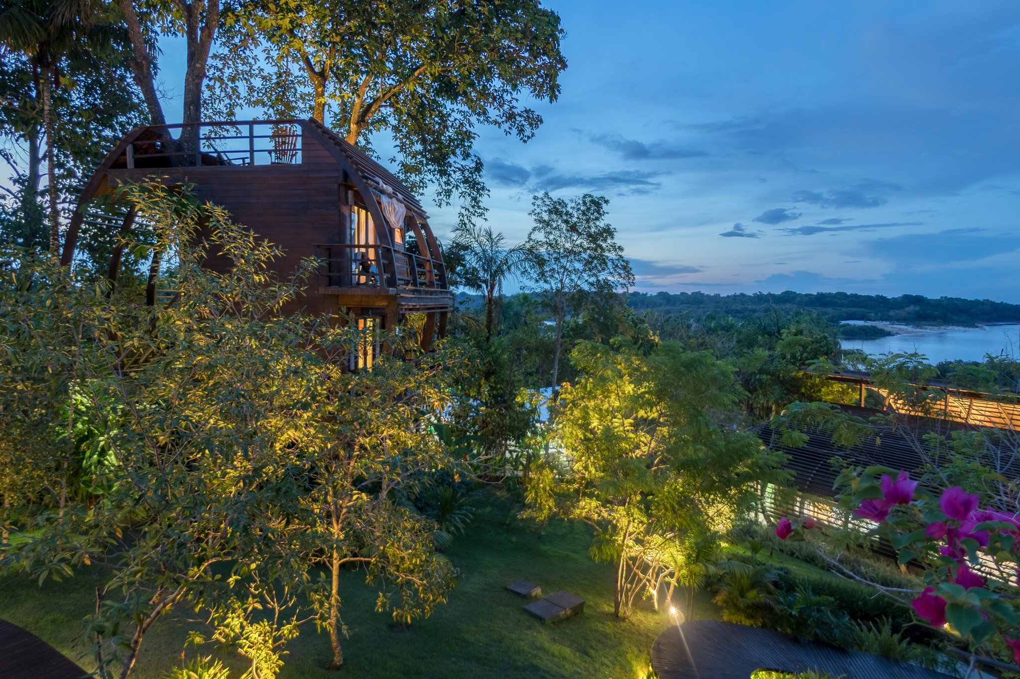 Illuminated curved wooden treehouse cabin at Mirante do Gavião rising above lush rainforest gardens at dusk, with the Rio Negro visible through the trees in the background