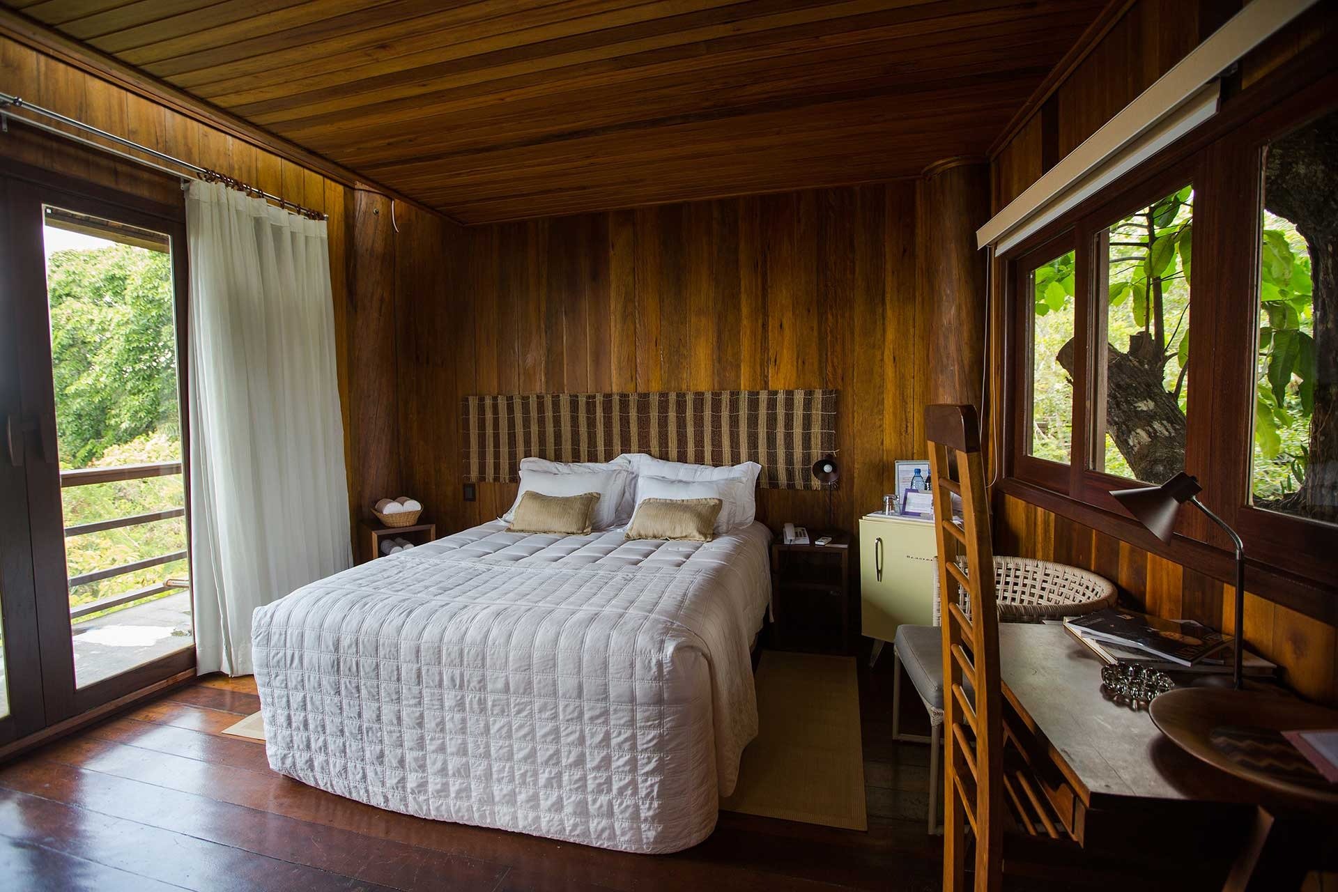 Wake up to the forest at your window — rustic warmth meets jungle serenity. Cozy double bedroom at Mirante do Gavião with warm timber-panelled walls, white quilted bedding, a woven headboard, and open glass doors revealing lush Amazon rainforest
