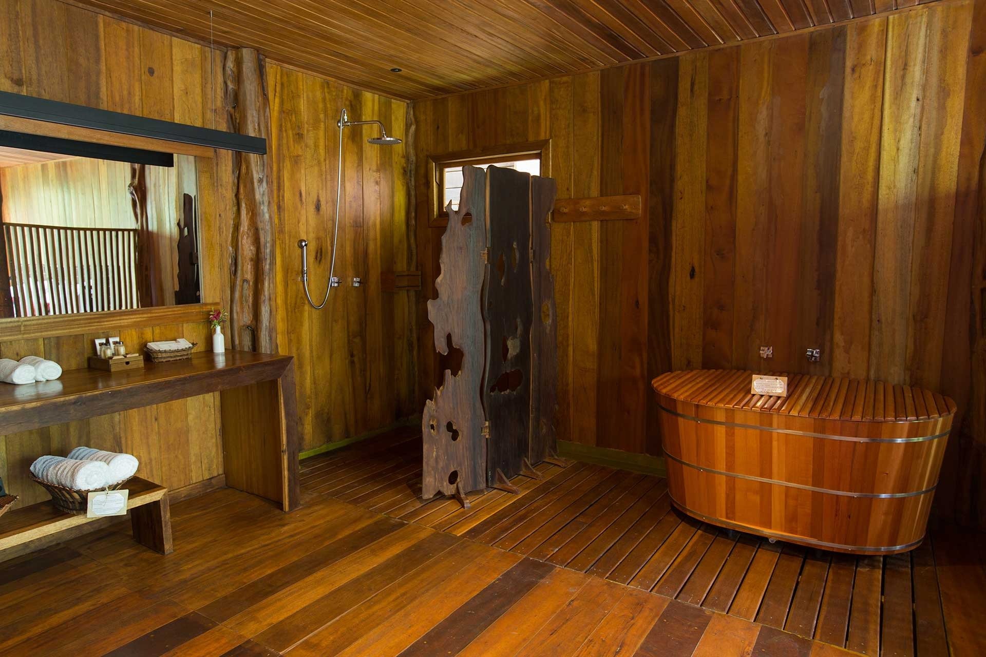 Raw timber, warm light, and a cedar soaking tub — comfort deep in the Amazon. Rustic all-wood bathroom at Mirante do Gavião featuring a round cedar soaking tub, rainfall shower, live-edge wooden divider, and neatly folded white towels on a timber bench