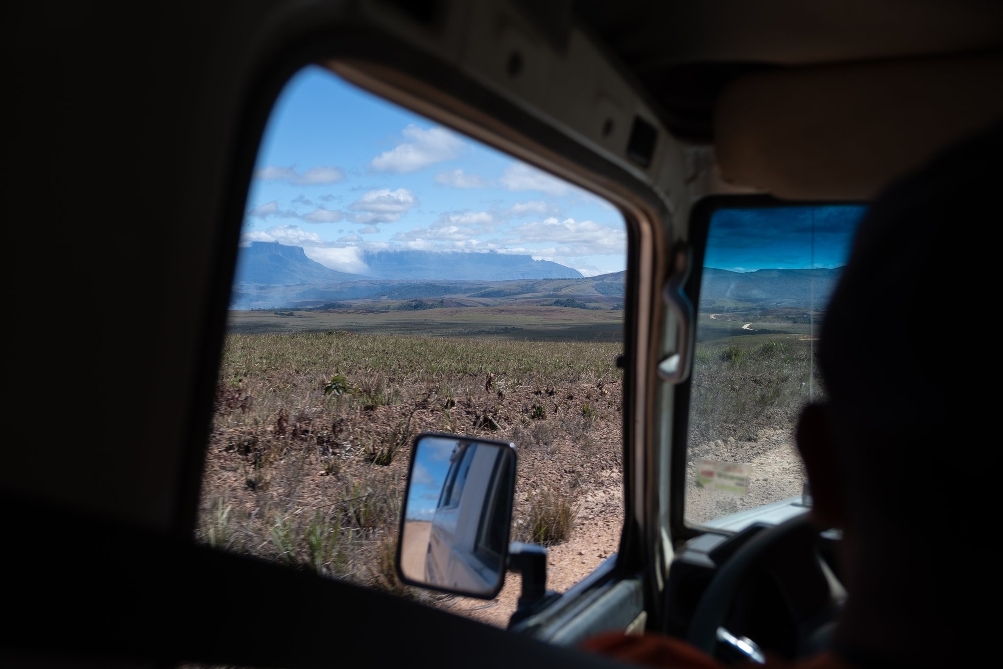 View through a 4x4 vehicle window of the vast Gran Sabana savanna with flat-topped tepui mountains on the horizon, framed by the wing mirror and a dusty dirt track — the approach to the Mount Roraima hike