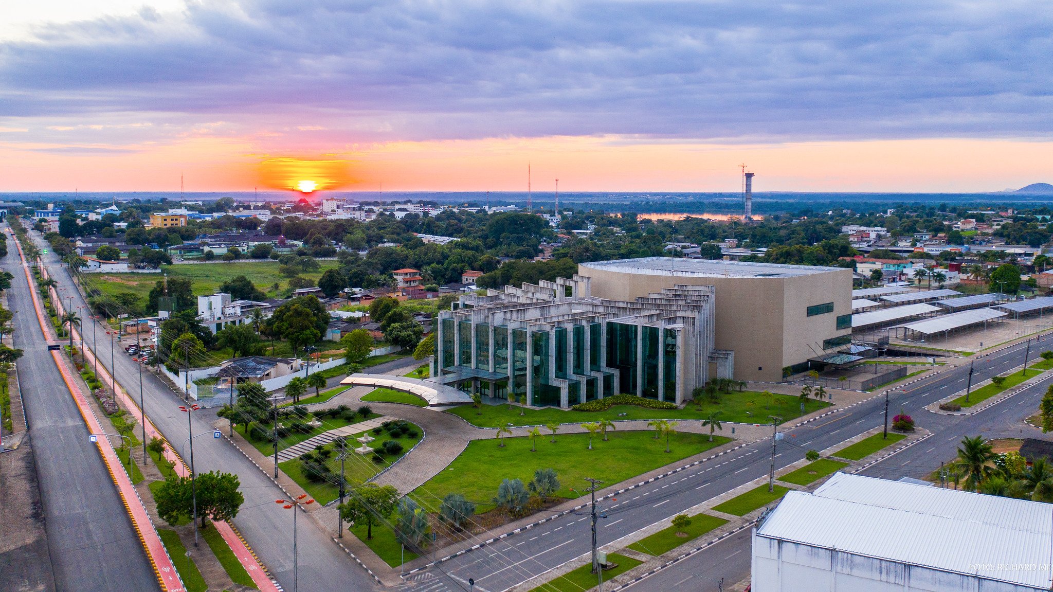 Aerial view of a Brazilian city at sunset, with a modernist glass-and-concrete civic building surrounded by green lawns and wide avenues, a glowing orange sun sinking toward the flat horizon — the gateway city for a Mount Roraima hike