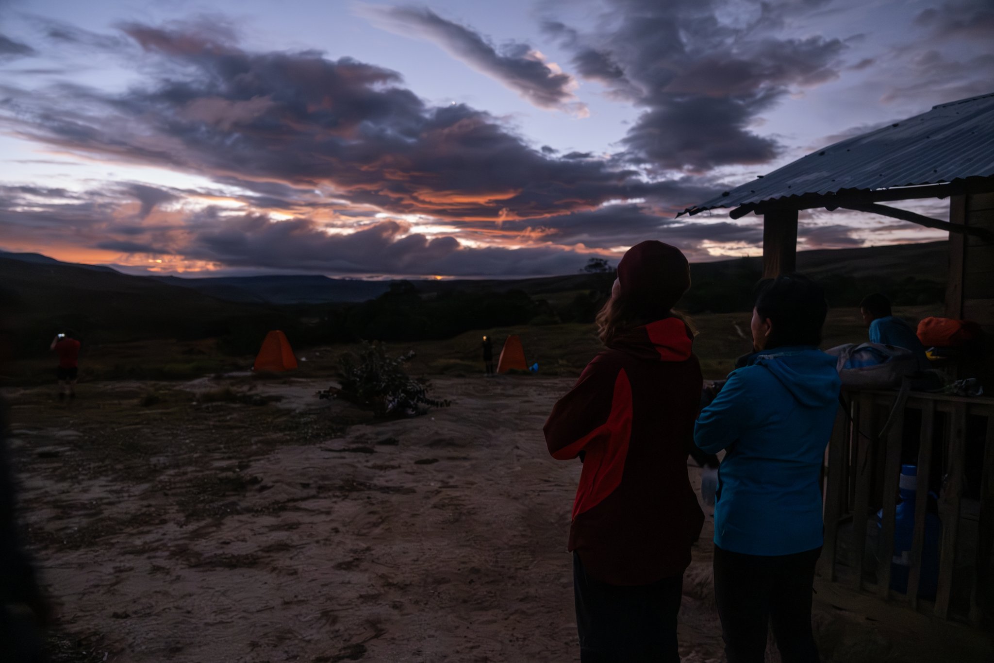 Trekkers in warm jackets watch a vivid orange and purple twilight sky from a remote campsite on the Mount Roraima hike, with orange dome tents glowing in the background