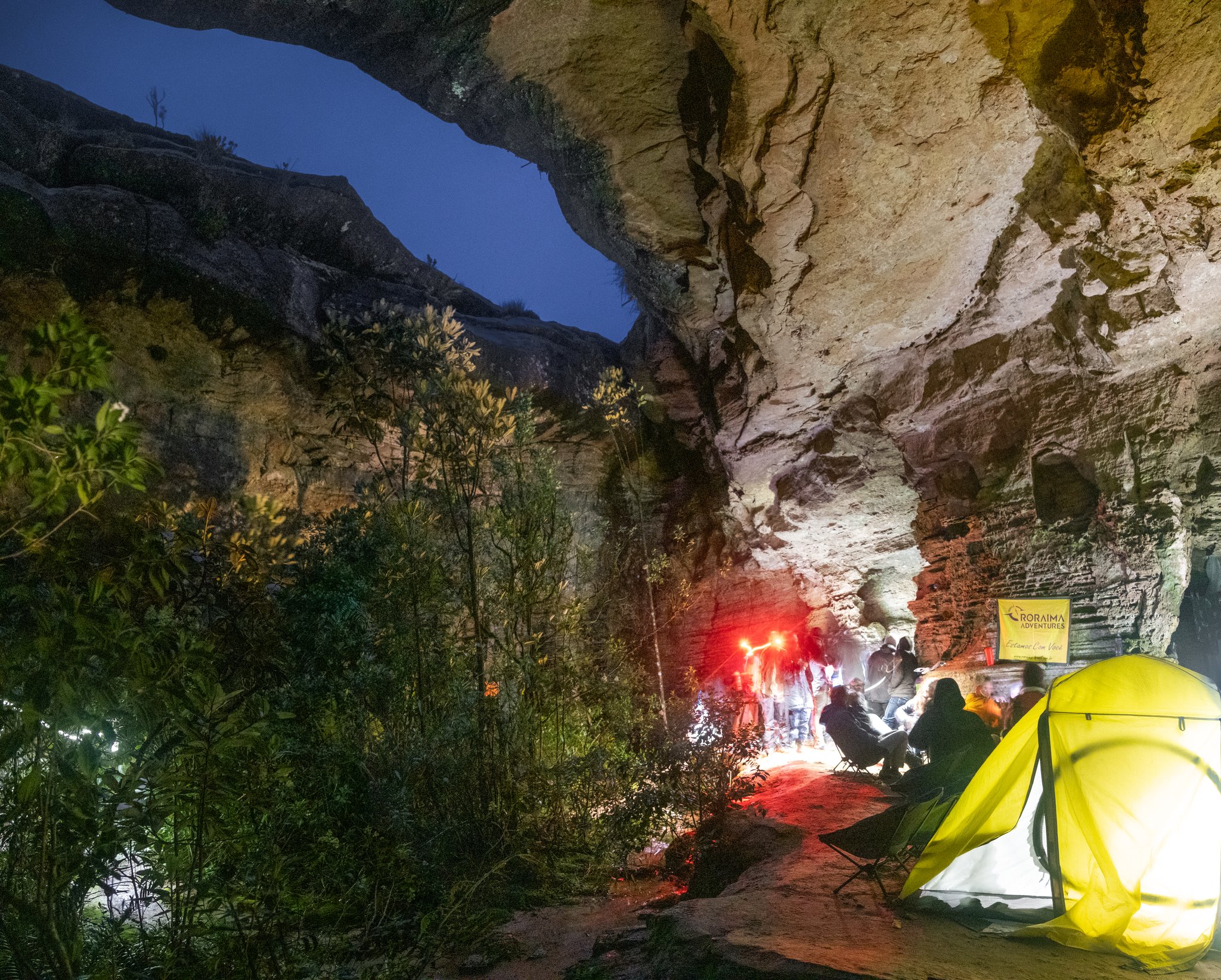 Trekkers camped under a massive sandstone rock overhang at night during the Mount Roraima hike, a glowing yellow tent lit from within beside a group gathered around red lights beneath ancient tepui walls
