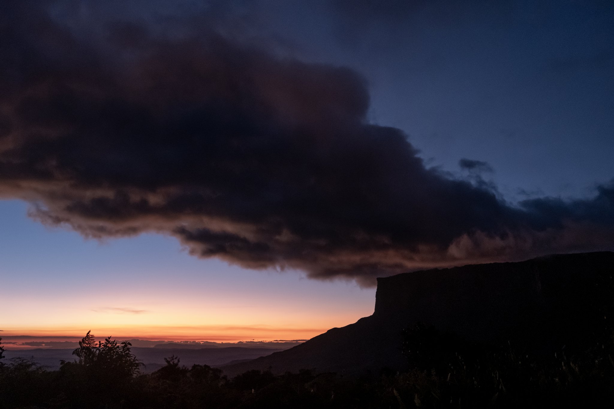 A massive dark storm cloud spreads over the silhouette of a flat-topped tepui during a vivid orange and deep blue sunset on the Mount Roraima hike
