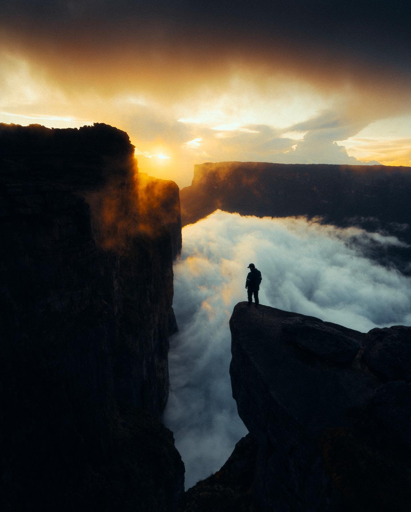 A lone hiker silhouetted on a rocky ledge during the Mount Roraima hike, gazing into a sea of clouds rolling between sheer tepui cliffs as a golden sunset blazes through the gap
