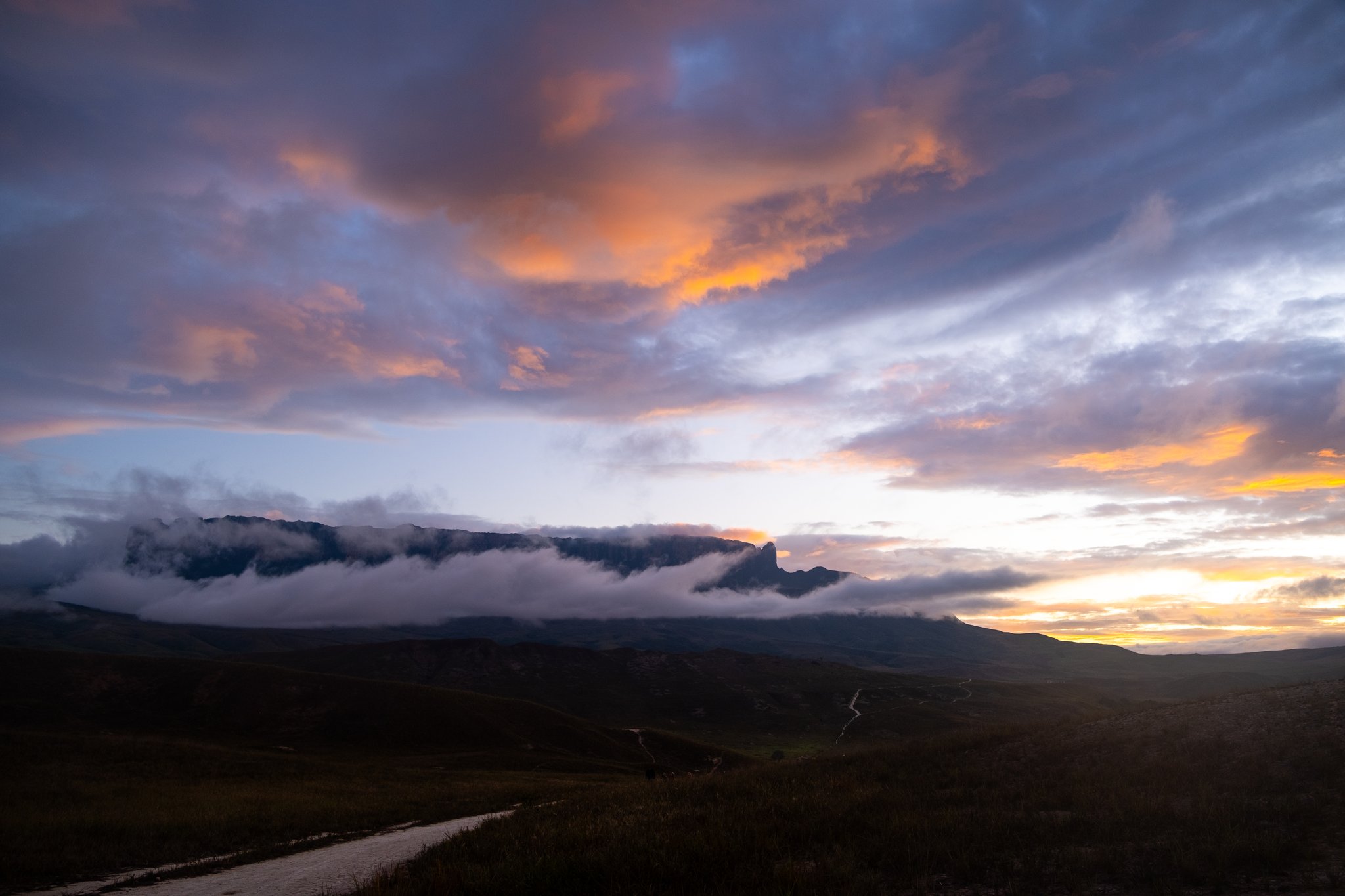 Dramatic sunset sky with orange and purple clouds billowing above a flat-topped tepui silhouette on the Mount Roraima hike, with a winding dirt track visible across the darkened valley below