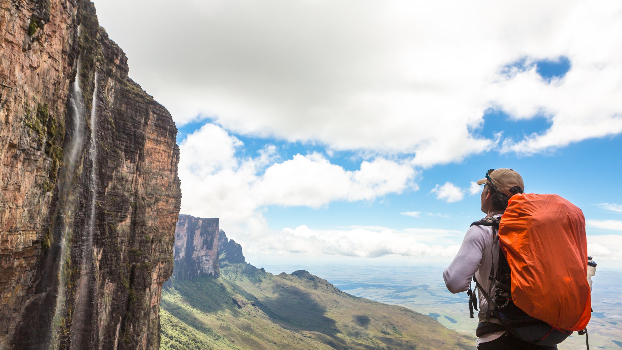 A hiker with an orange pack gazes up at a sheer sandstone cliff with a thin waterfall cascading down it during the Mount Roraima hike, with a vast savanna plain stretching to the horizon below