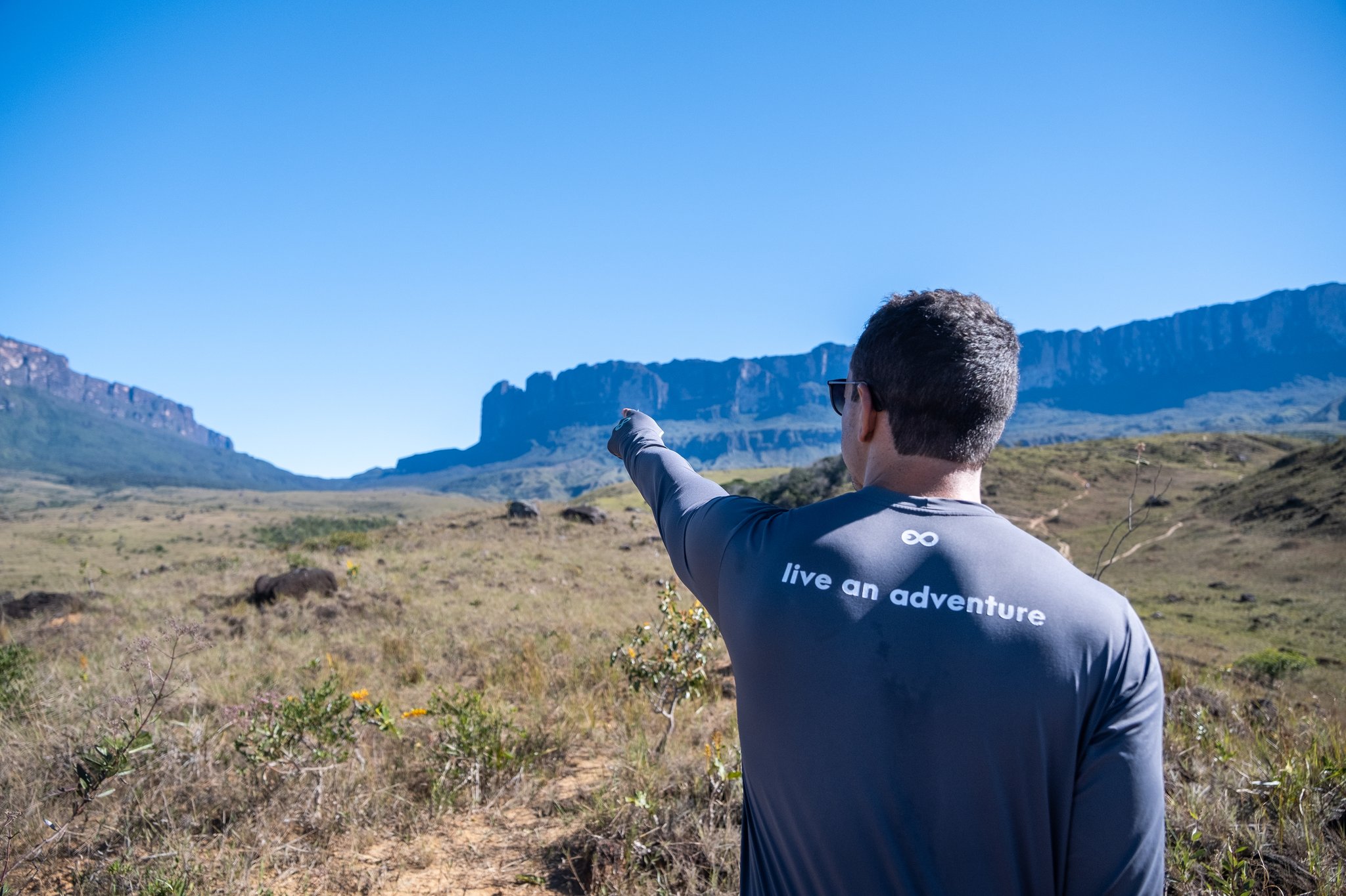 A trekker in a 'live an adventure' shirt pointing toward the dramatic flat-topped cliffs of Mount Roraima across an open savanna under a clear blue sky