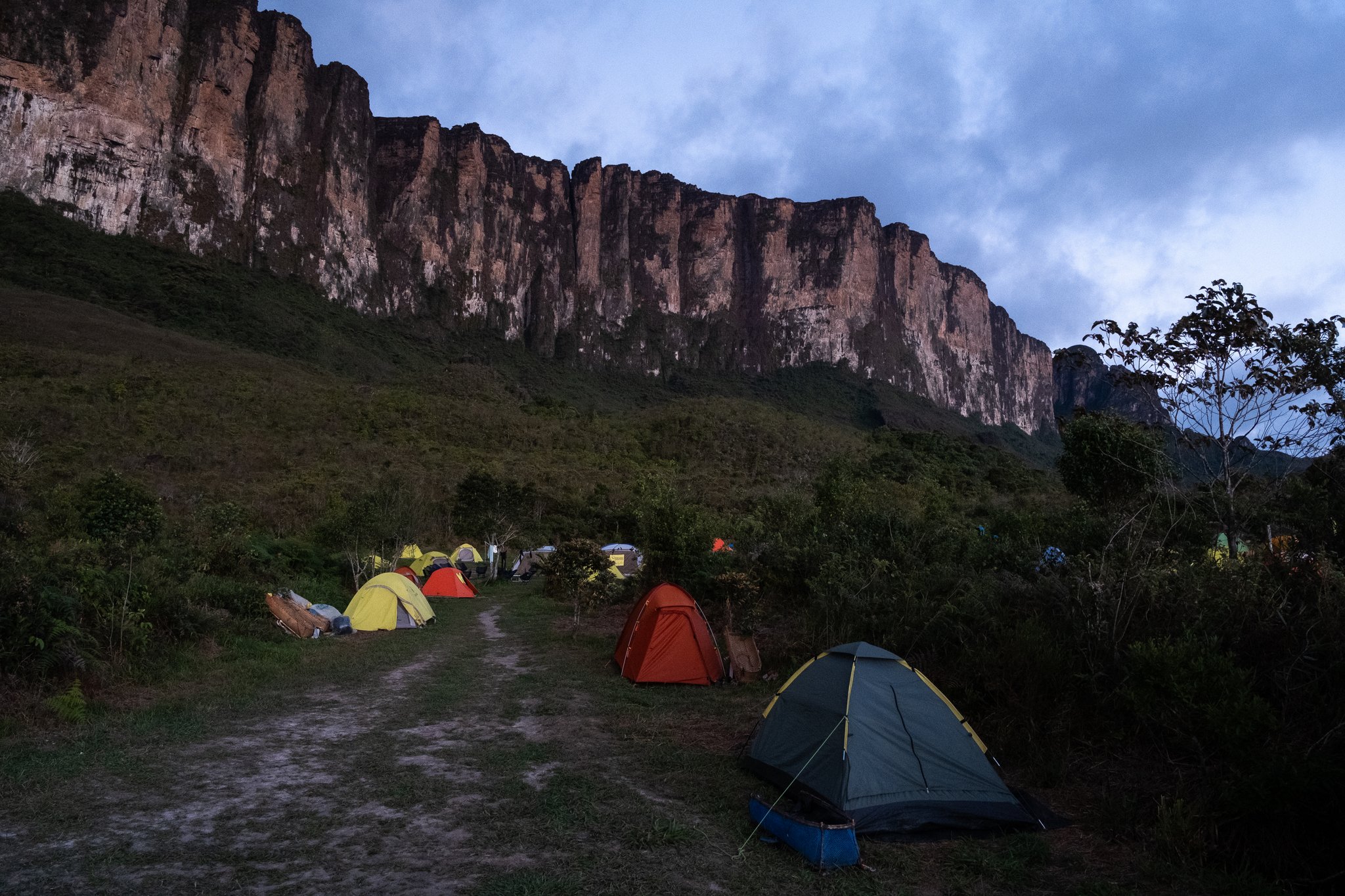 Colorful tents pitched at a base camp on the Mount Roraima trek, with towering ancient sandstone cliffs glowing red under a moody twilight sky