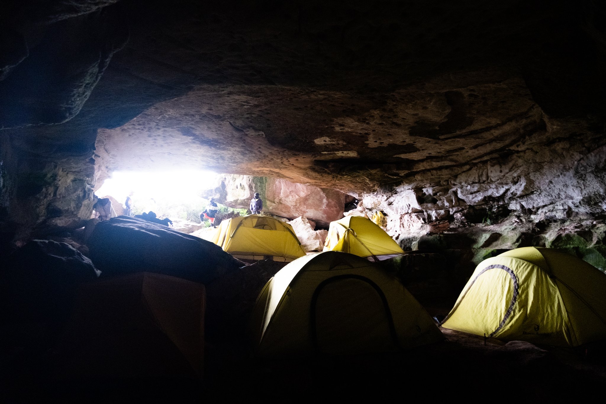 Yellow dome tents pitched inside a rocky cave shelter during a Mount Roraima trek, with daylight visible through the low cave entrance and trekkers silhouetted outside