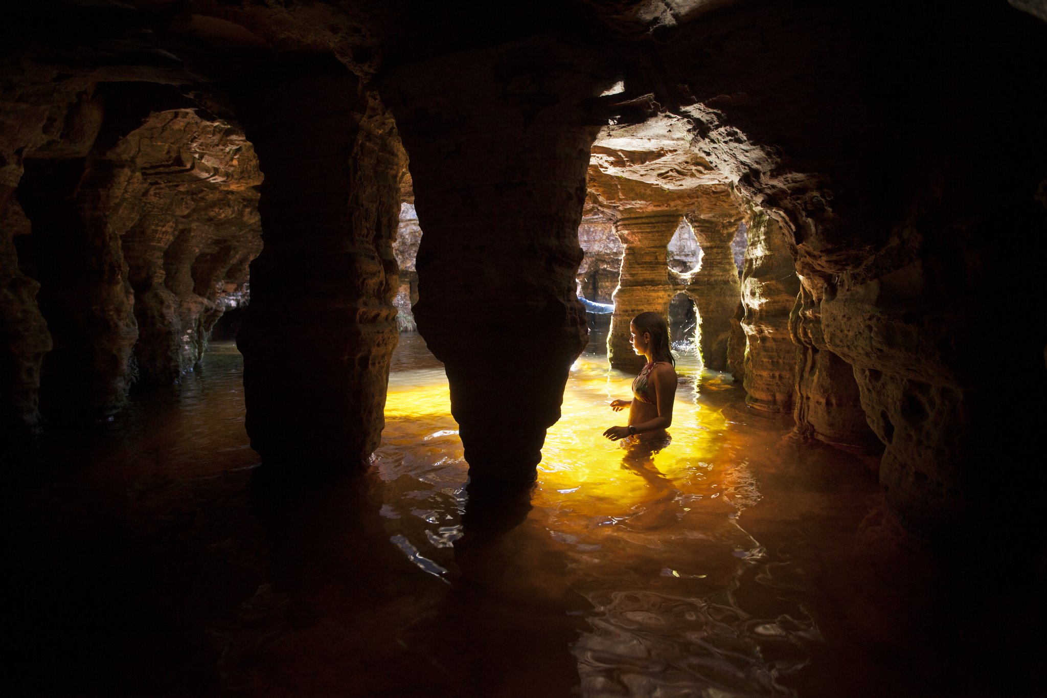 A woman wades waist-deep through a glowing amber underground pool inside a sculpted sandstone cave on the Mount Roraima trek, light filtering through eroded rock arches
