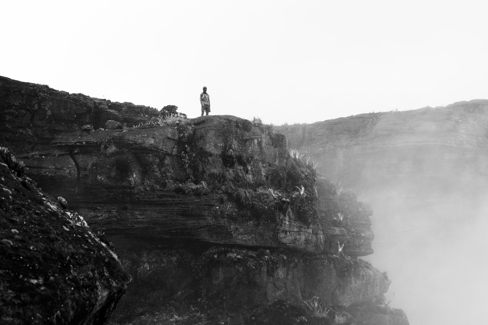 A lone hiker stands on the edge of a moss-covered sandstone cliff during a Mount Roraima trek, mist rising from the canyon below in a dramatic black-and-white scene