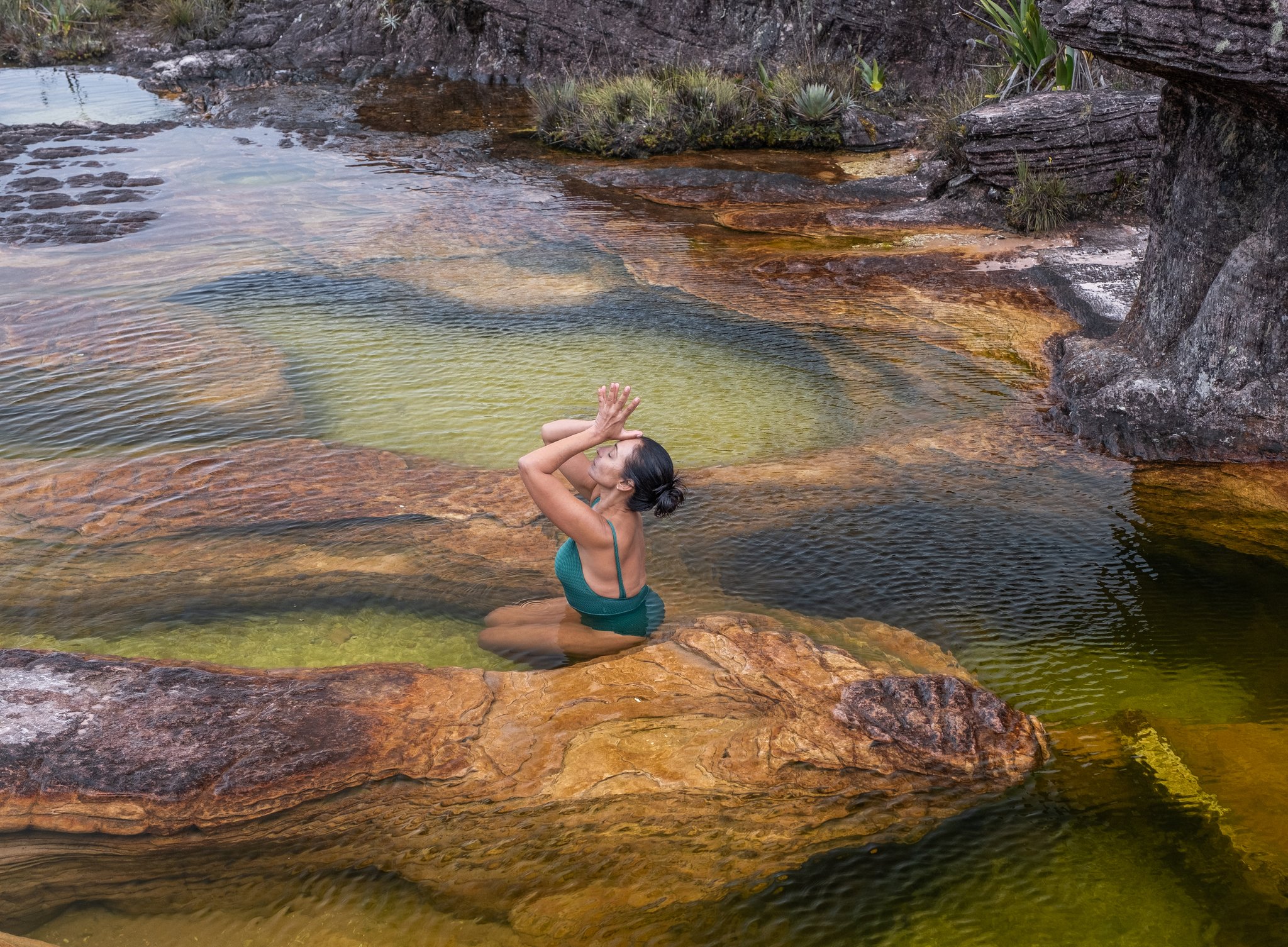 A woman in a teal swimsuit kneels in a shallow natural pool on the Mount Roraima trek, hands raised in a meditative pose above rust-coloured tepui rock carved by ancient water erosion
