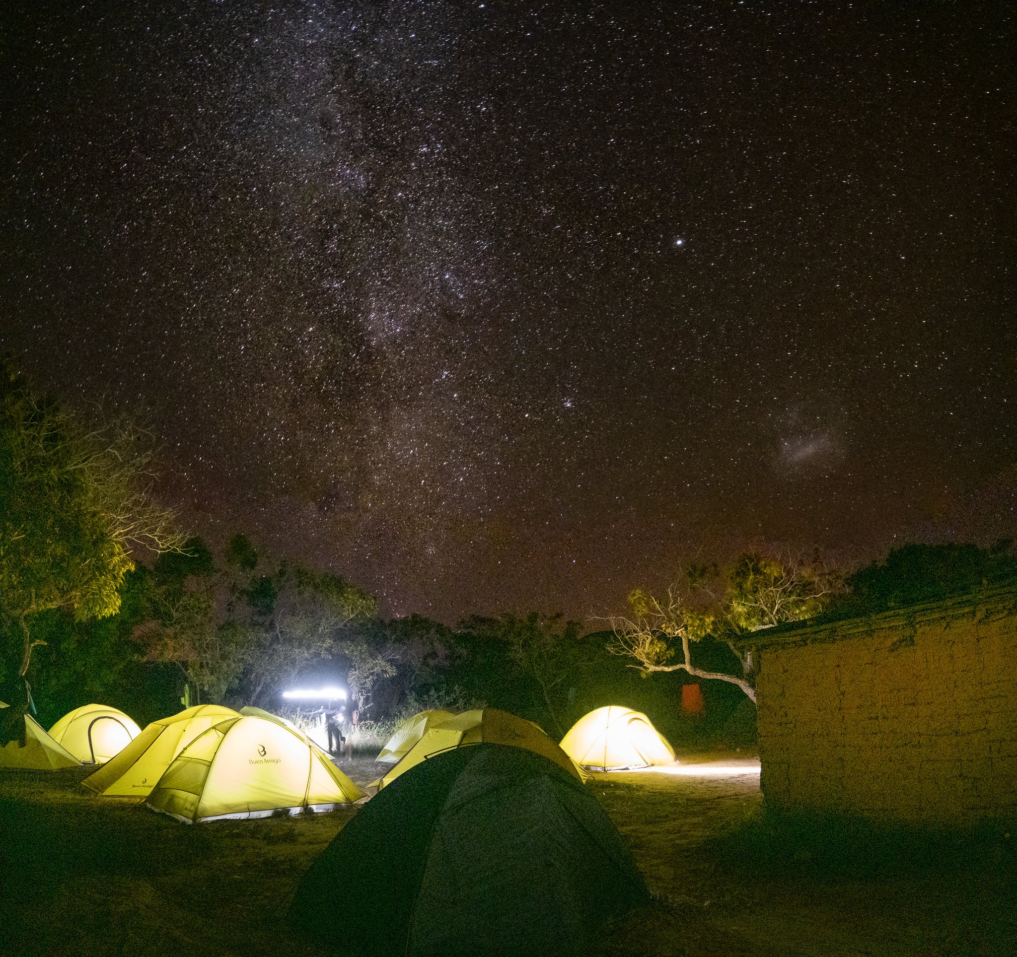 Glowing yellow tents clustered at a wilderness campsite on the Mount Roraima trek beneath a brilliant Milky Way sky, with a lone figure standing in the lamplight