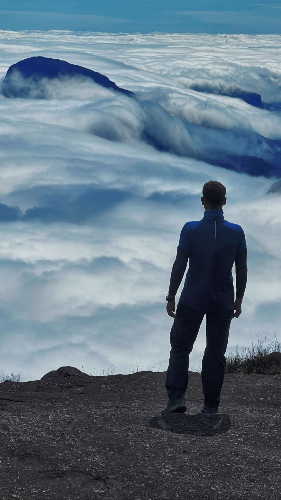 A hiker stands on the rocky summit plateau of the Mount Roraima trek, gazing out over a vast sea of clouds that swallows the surrounding tepuis below