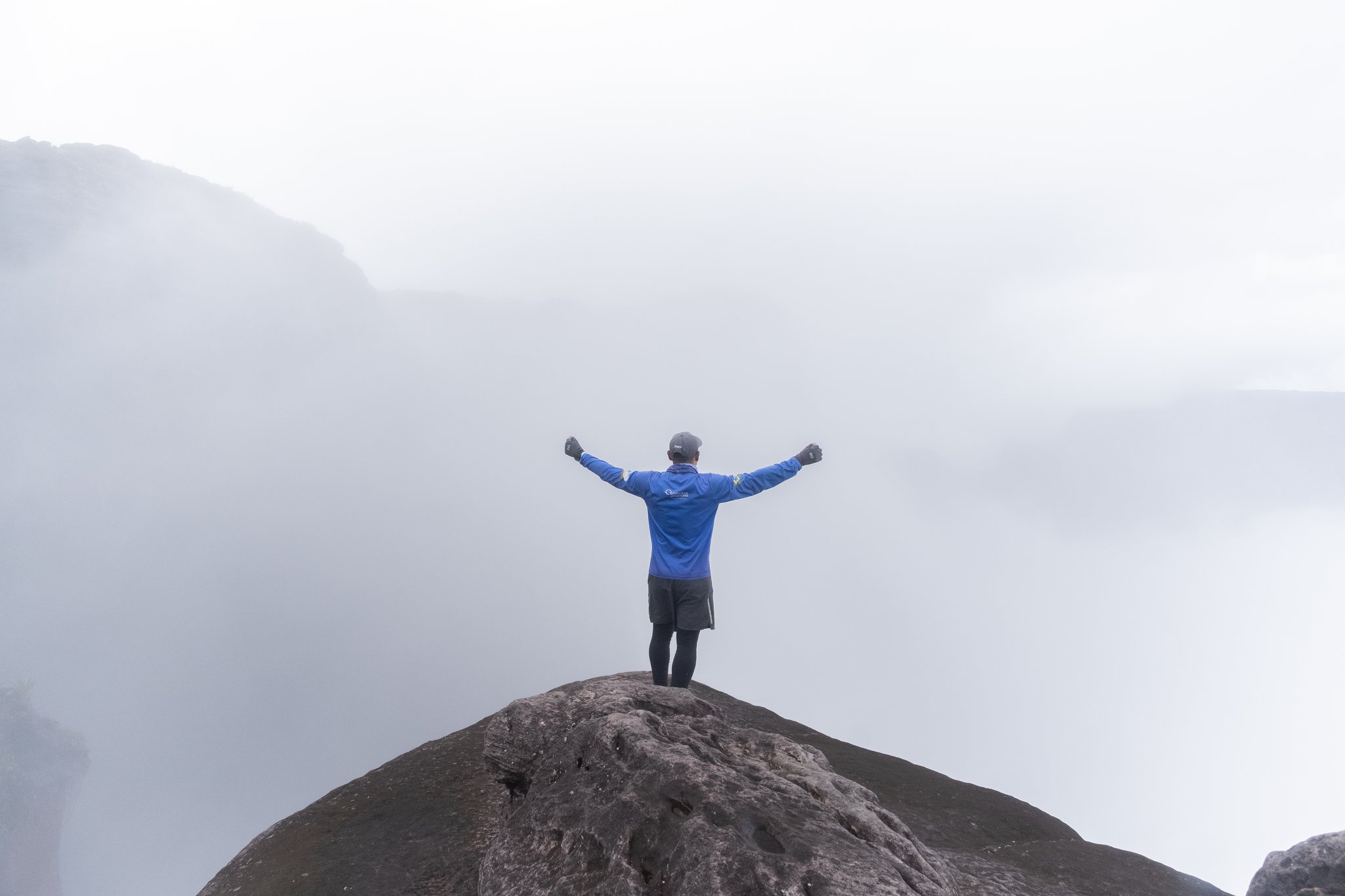 A hiker in a blue jacket stands arms wide open on a bare rocky outcrop during the Mount Roraima trek, surrounded by thick swirling mist and steep cliff faces