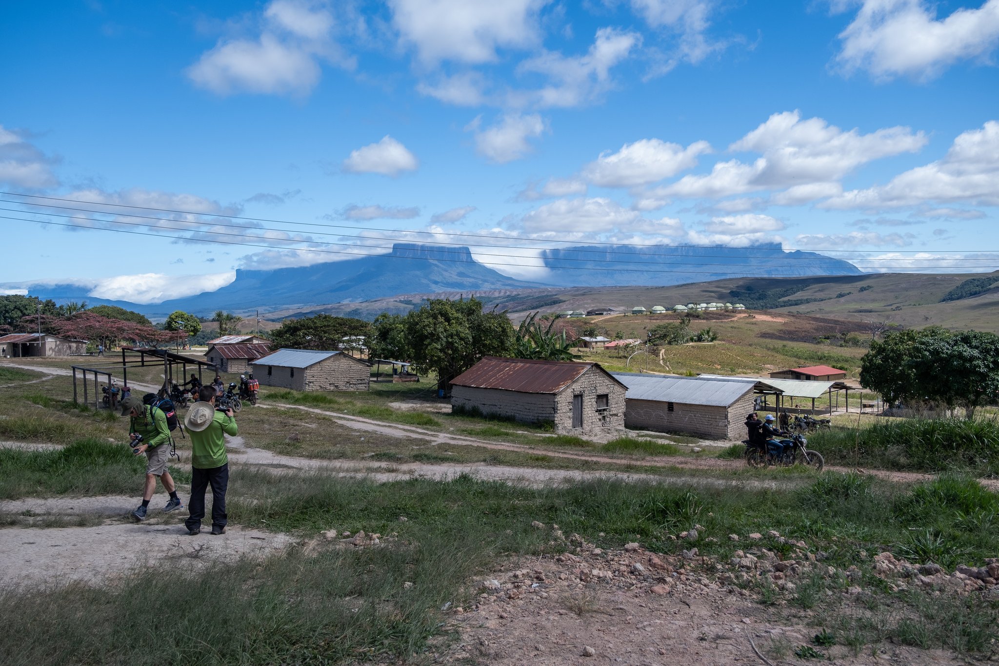 Two photographers capture the view from a dirt path above a small village on the Mount Roraima trek, with a flat-topped tepui rising dramatically on the horizon under a blue sky with white clouds