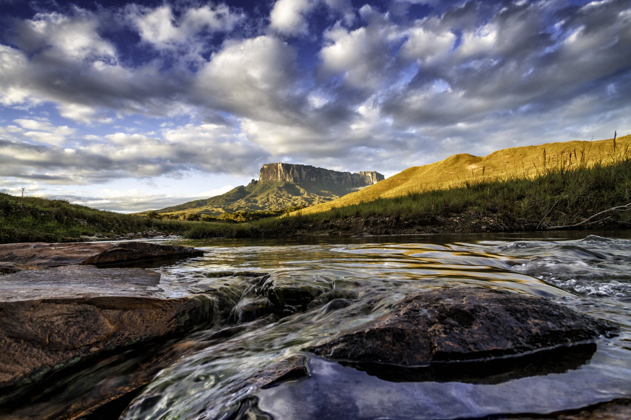 A shallow rocky river flows toward the flat-topped summit of Mount Roraima under a dramatic cloudscape on the Mount Roraima trek