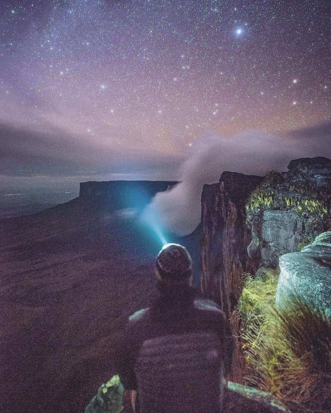 A lone hiker wearing a headlamp stands at the edge of a sheer tepui cliff on the Mount Roraima trek, gazing across a sea of mist toward a distant flat-topped plateau beneath a vast, star-filled sky