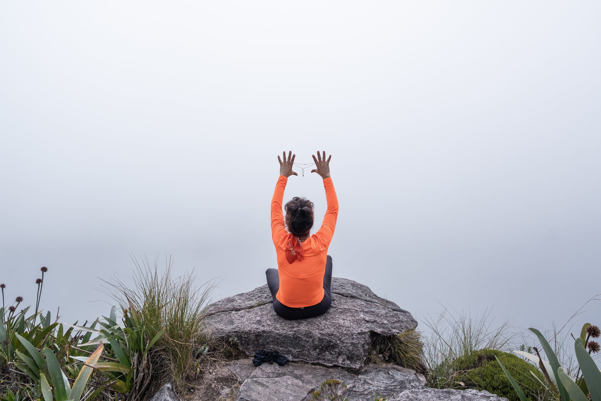 A woman in an orange top sits cross-legged on a mossy summit rock during the Mount Roraima trek, arms raised toward a sky swallowed by dense white cloud, surrounded by highland bromeliads