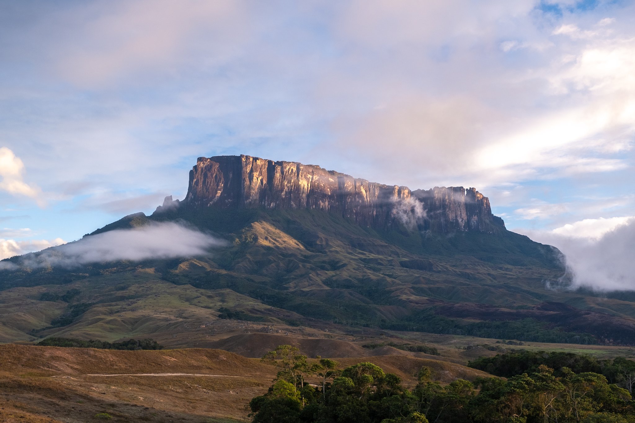 The flat-topped sandstone massif of a tepui rises above rolling savannah grasslands during a Mount Roraima trek, with low clouds drifting around its sheer vertical cliffs in warm morning light