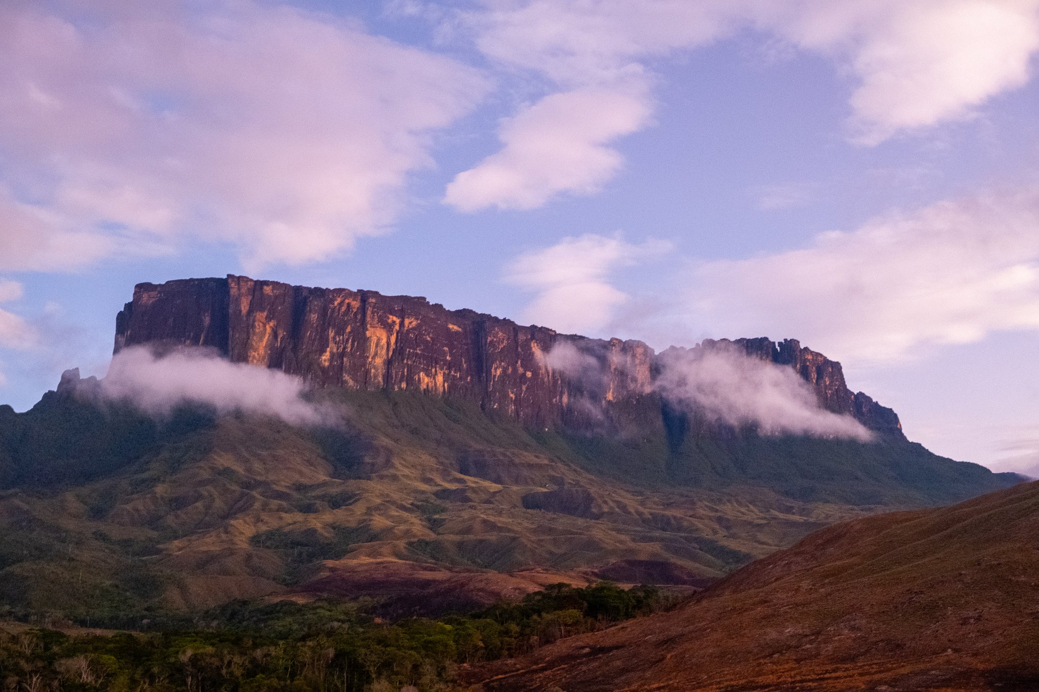 A flat-topped tepui rises above rolling green savannah on the Mount Roraima trek, its sheer sandstone cliffs glowing amber in low light while mist clings to the base of the plateau
