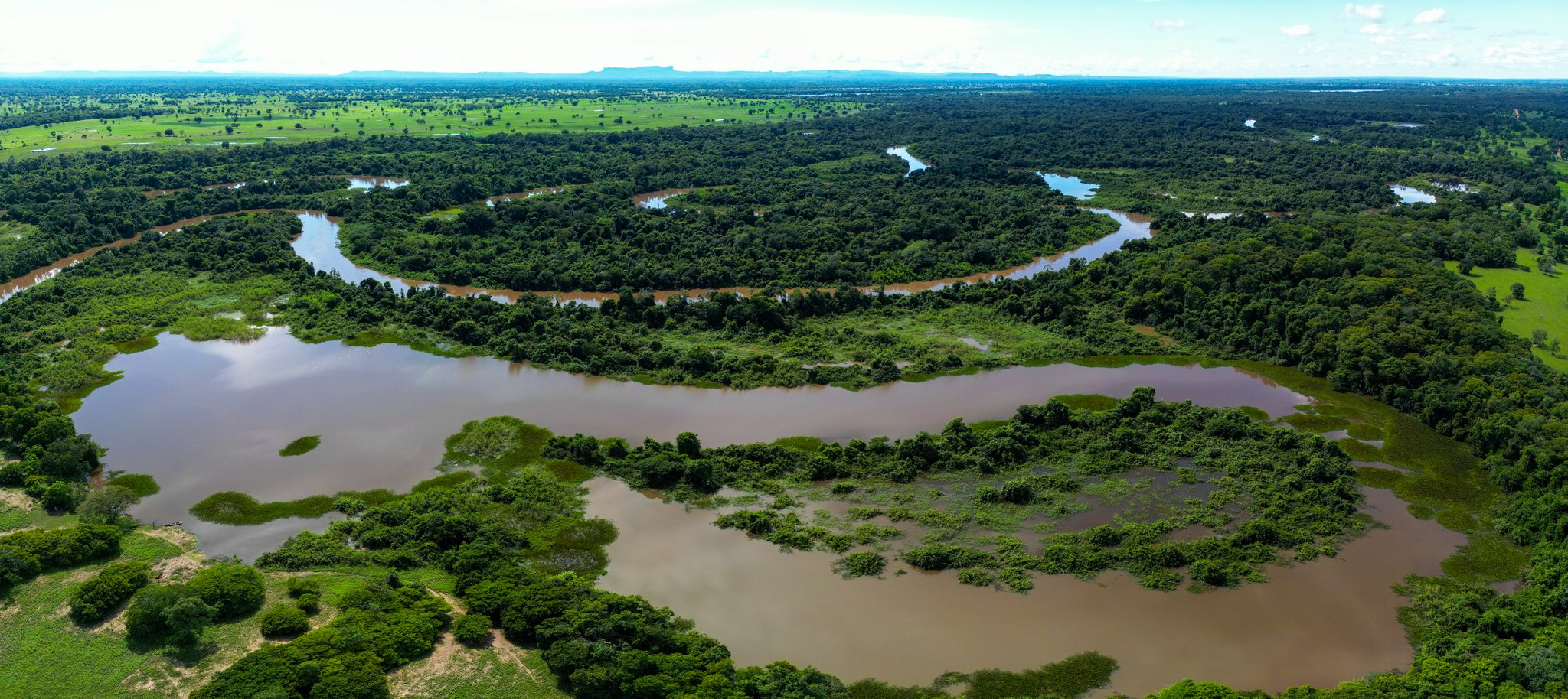 Endless water, endless green — the Pantanal sprawls to the horizon. Aerial panorama of a broad meandering river winding through dense tropical forest and flooded plains on a Pantanal Bonito tour, with distant mountains on the horizon