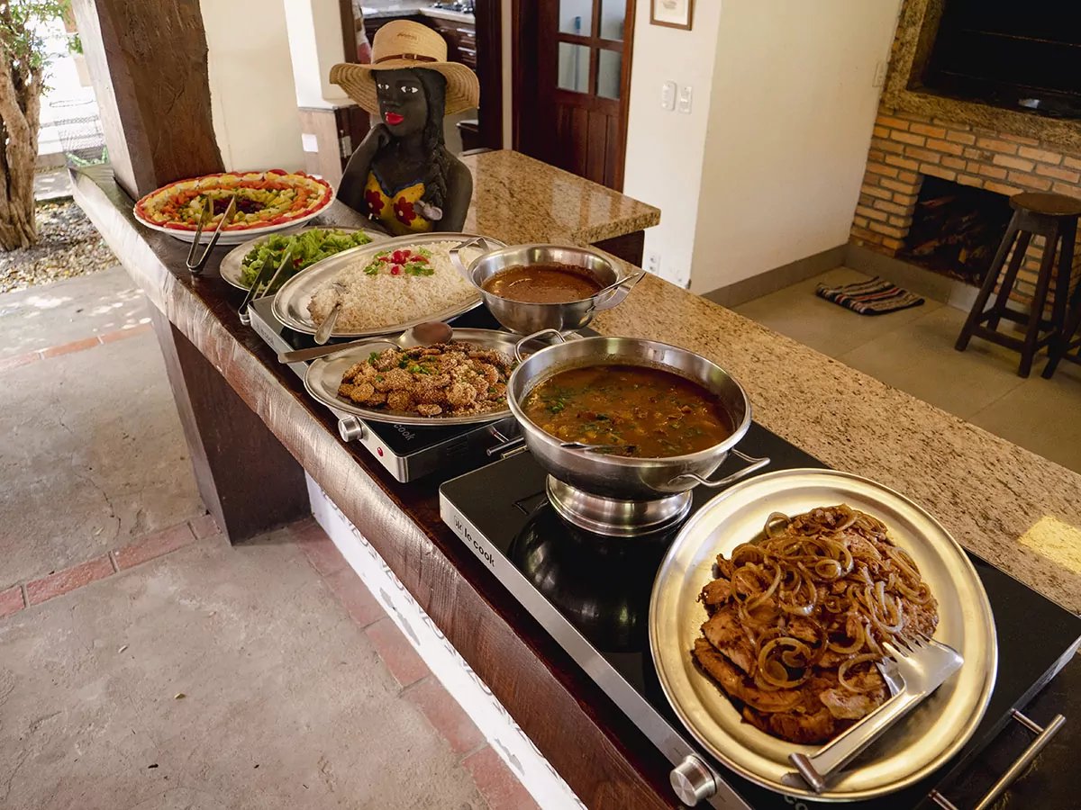 Hearty stews, seasoned meats and fresh salads — lodge cooking that refuels you for the next adventure. A granite-topped buffet counter laid with steaming pans of stew, seasoned meat with caramelised onions, rice, fried bites, salads and a colourful fresh-fruit platter — hearty Brazilian lodge cooking on a Pantanal Bonito tour