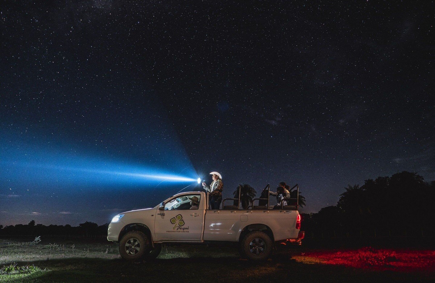 Spotlight on — the night safari begins under a sky thick with stars. A guide standing in the back of a Pantanal lodge truck sweeps a powerful spotlight across a star-filled night sky during a Pantanal Bonito tour night safari