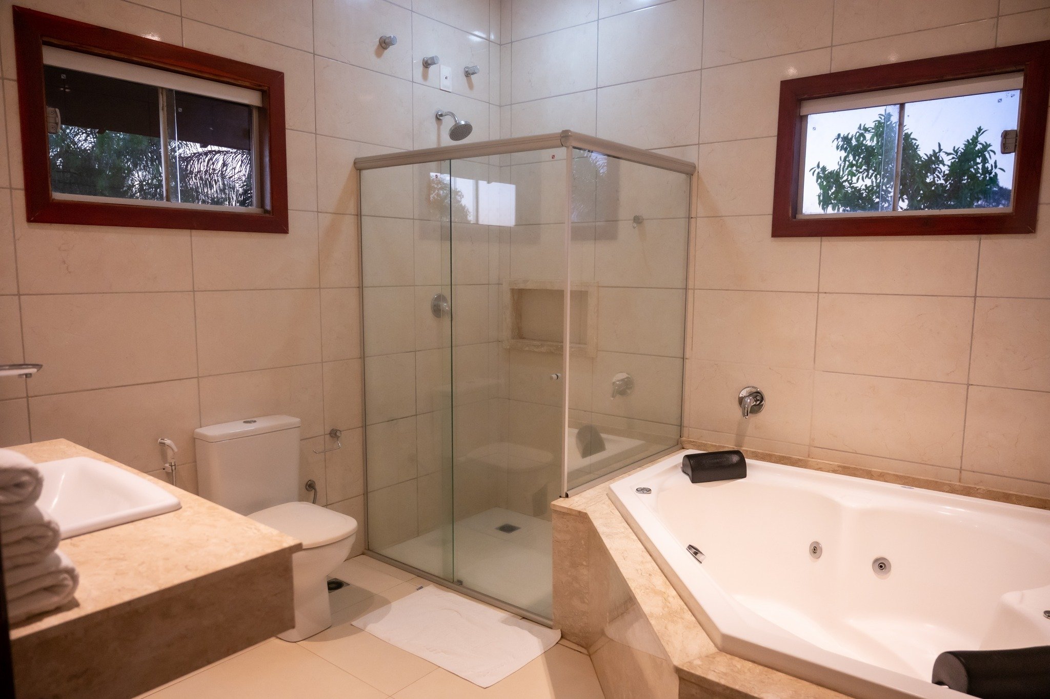 Clean tile, a corner jacuzzi, and leafy views through the window — comfort well earned after a day on the river. Spacious en-suite bathroom at a Pantanal jaguar budget safari lodge, featuring a glass-enclosed shower, corner jacuzzi tub, marble-surround vanity, and windows overlooking tropical greenery