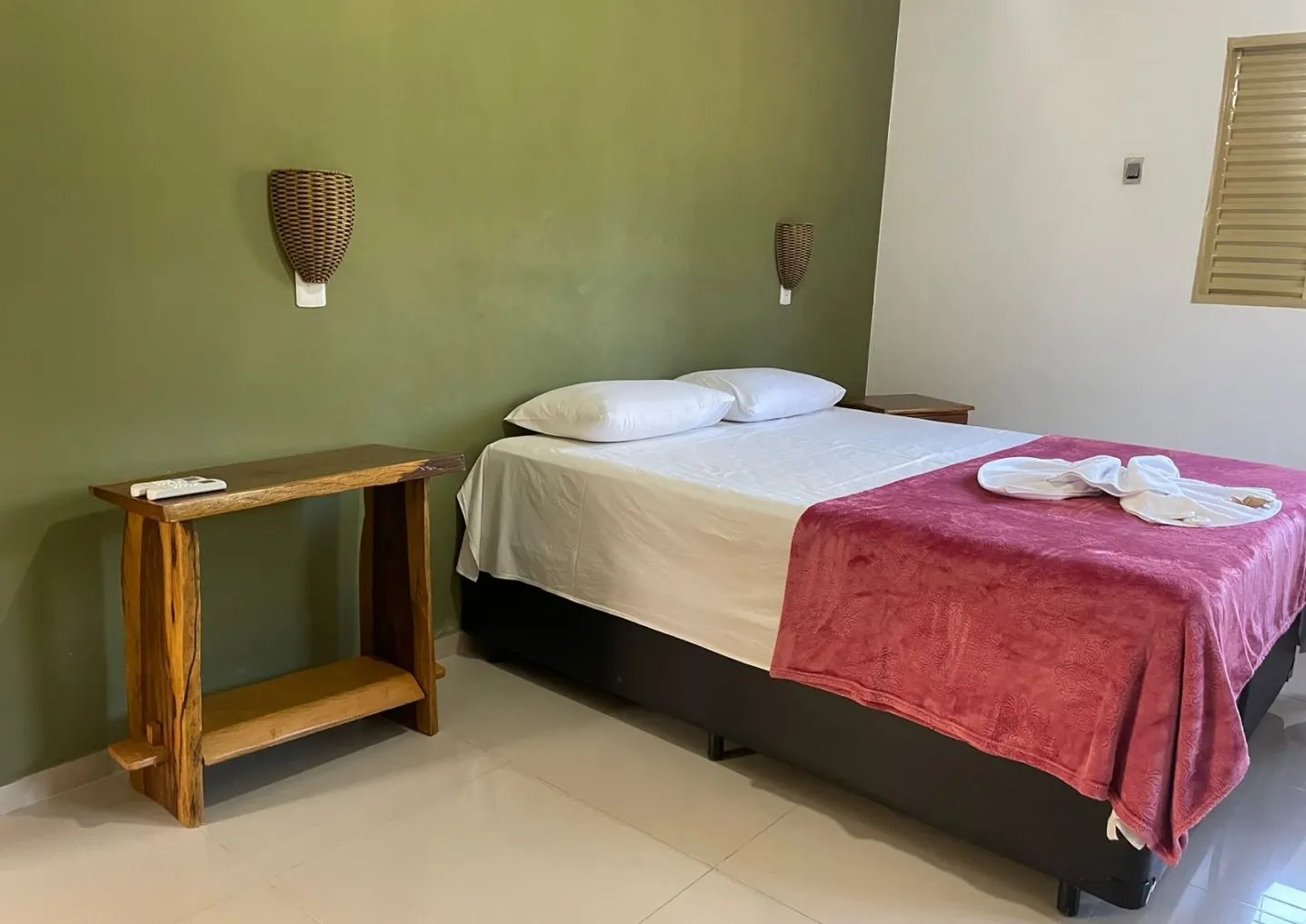 Rest well before the river — simple, comfortable rooms ready for safari days. A clean double room on a pantanal jaguar budget safari, featuring a double bed with white linens and a red throw, wicker wall sconces on a sage-green wall, and a rustic live-edge wooden side table