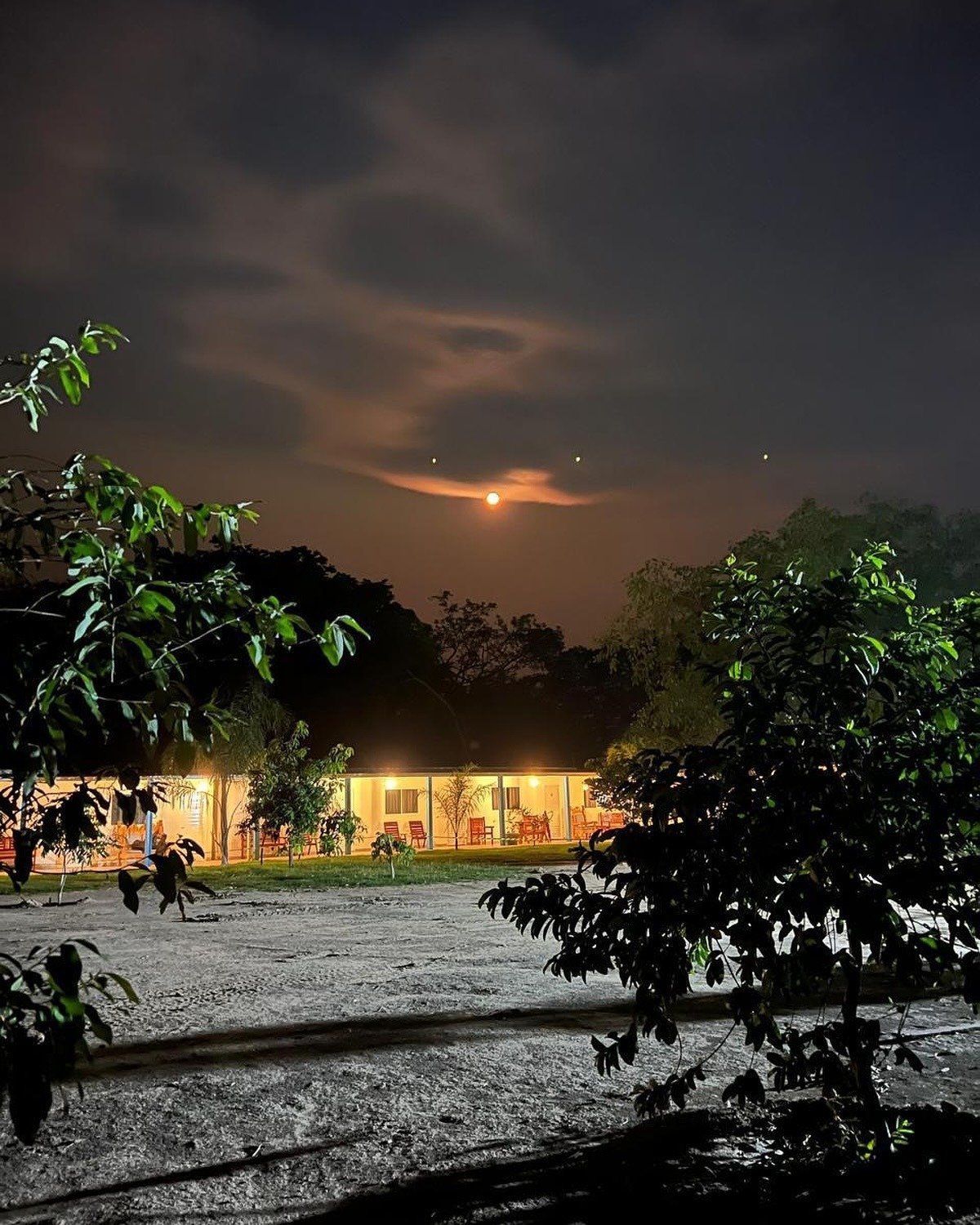 Full moon over the lodge — the Pantanal night begins. A warmly lit lodge glows against a dramatic night sky on a pantanal jaguar budget safari, with a full moon rising through dark clouds above tropical trees