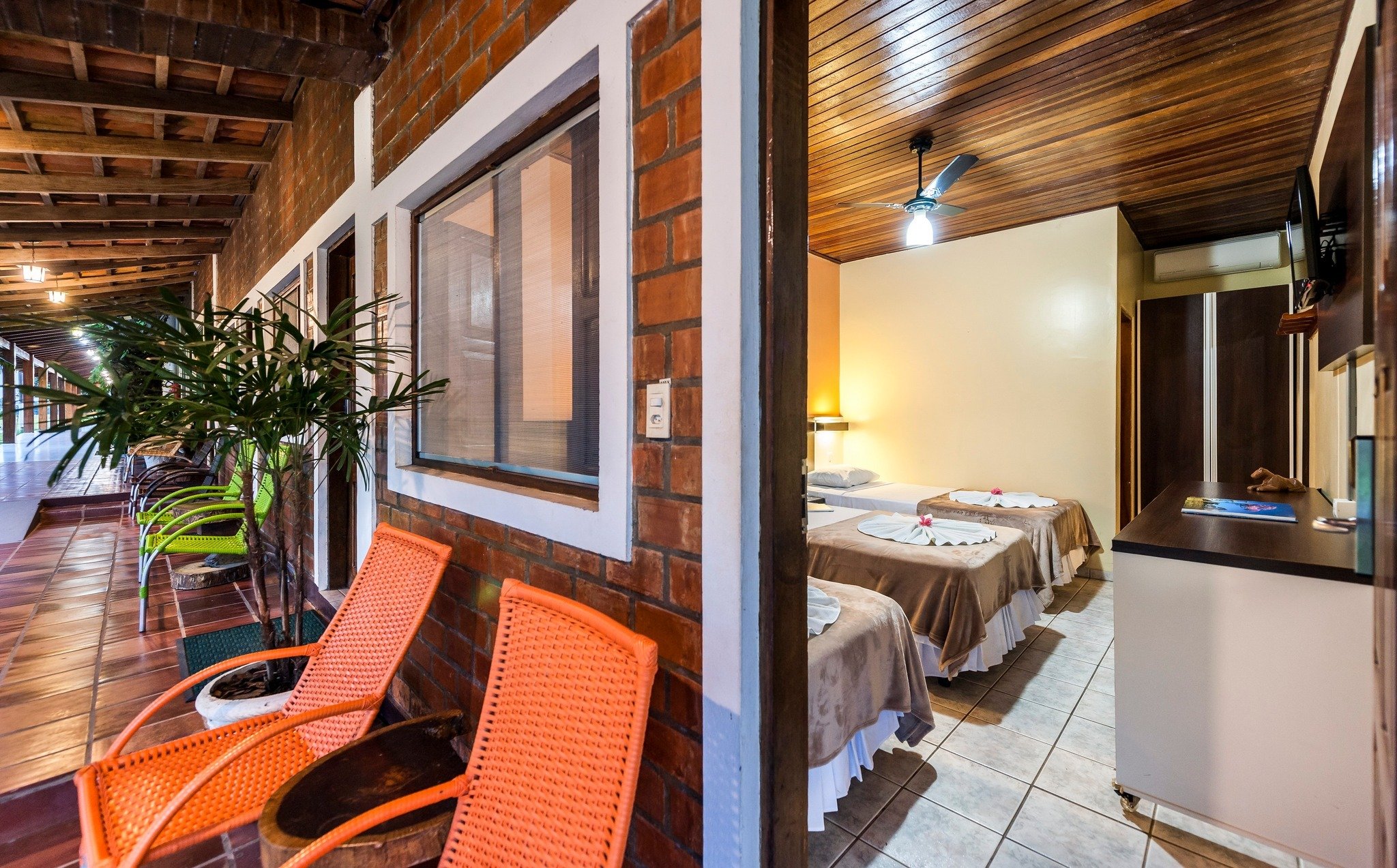 Rattan chairs on the veranda, flower-trimmed beds just beyond the door — comfort kept simple. Covered brick veranda with orange rattan chairs leading to a twin bedroom with ceiling fan and flower-decorated beds, typical of a pantanal jaguar budget safari lodge