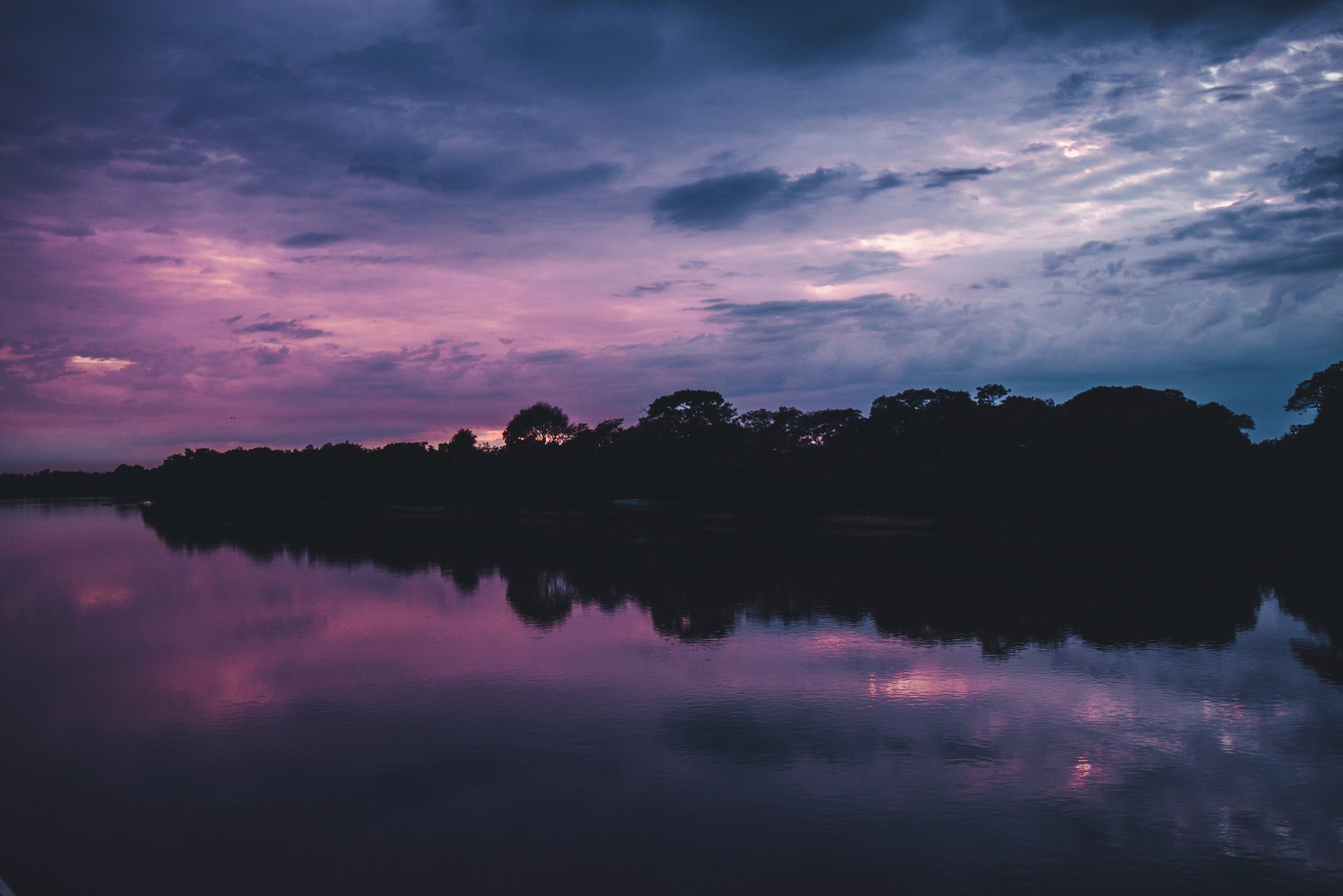 Pink fades to violet as the Pantanal river mirrors the sky. A vivid purple and pink twilight sky reflected on a still river, with a dark treeline silhouetted along the far bank on a pantanal jaguar budget safari