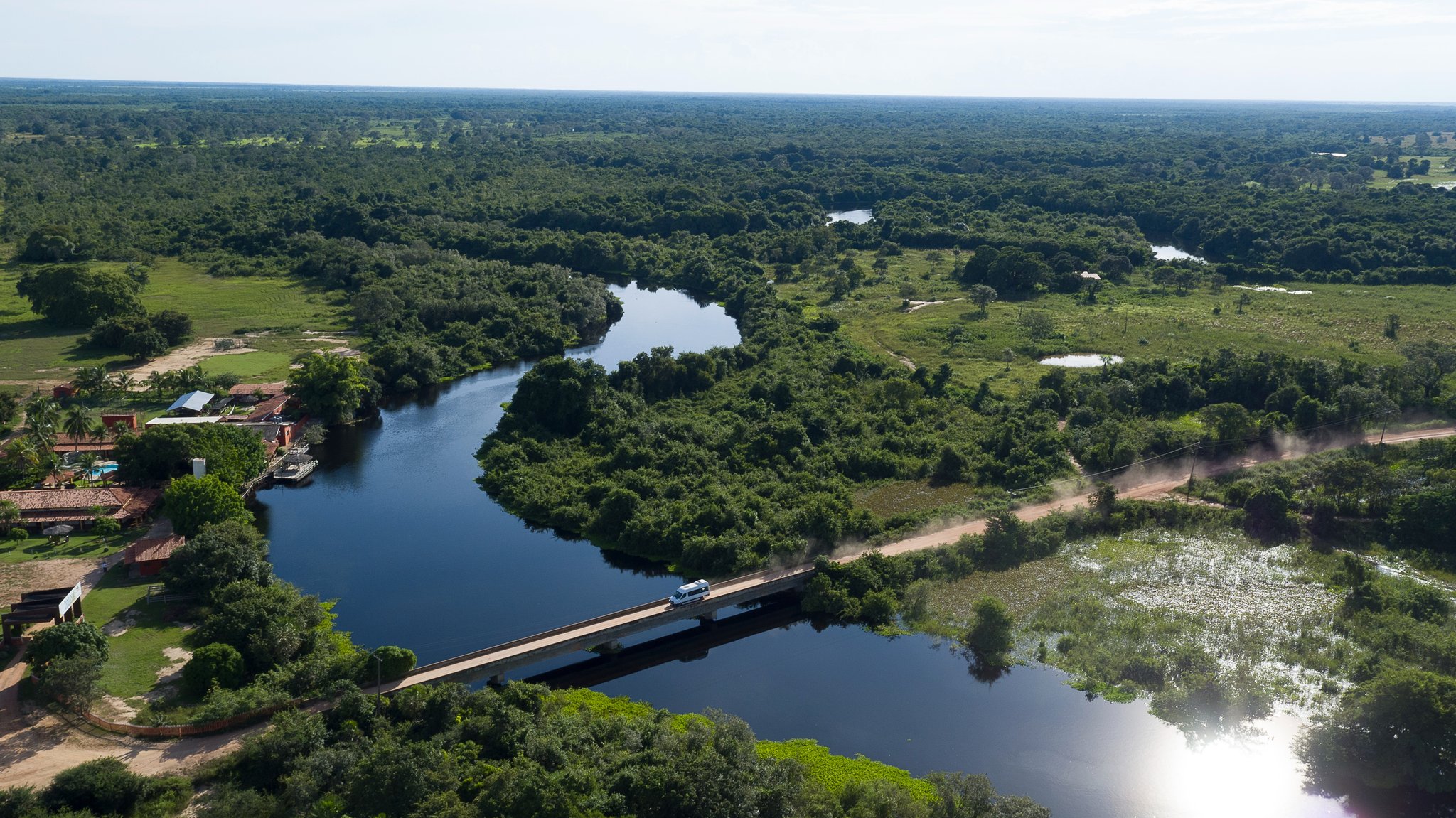 A single bridge, a winding river, and the whole wild Pantanal ahead. Aerial view of a white van crossing a concrete bridge over a winding river surrounded by dense Pantanal wetland vegetation and a rustic riverside lodge, the gateway to a pantanal jaguar budget safari