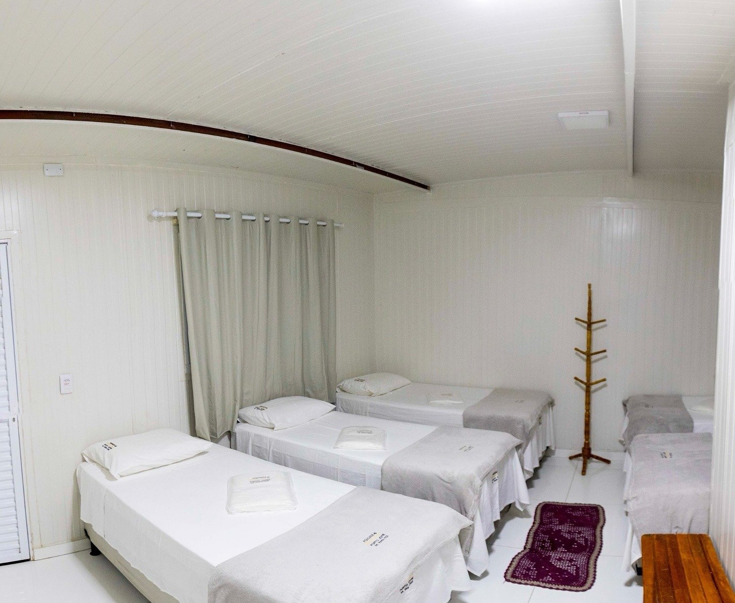 Fresh linens, four beds, and everything you need — comfort kept simple so the budget goes toward the wildlife. Clean shared dormitory room with four single beds dressed in white linen, a wooden coat stand, and a curtained window at a Pantanal jaguar budget safari lodge