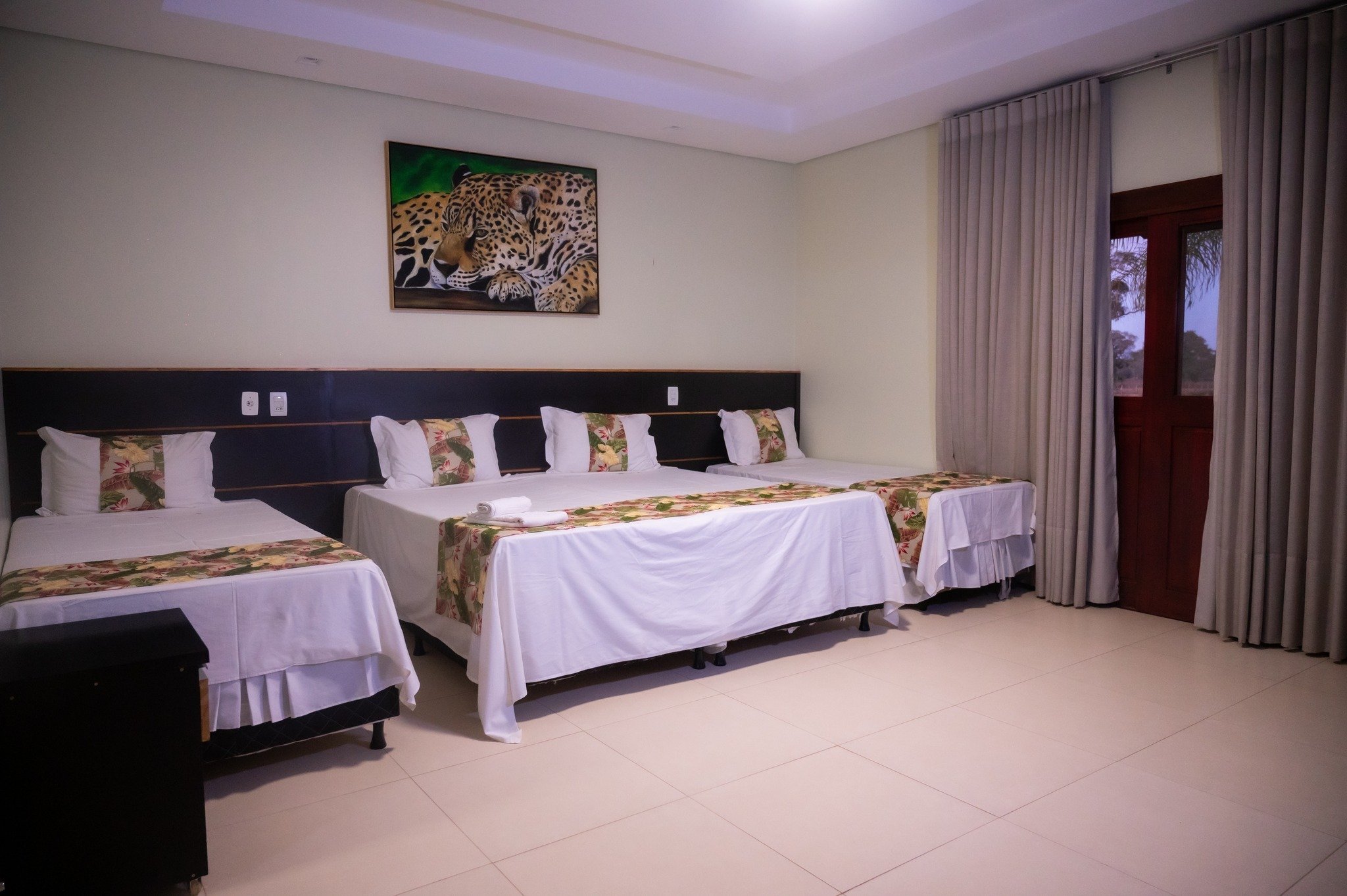 Three beds, crisp white linens, and a jaguar watching over you. Spacious triple-bed guest room on a pantanal jaguar budget safari, with white linens, tropical-print bed runners, and a jaguar painting above the dark headboard