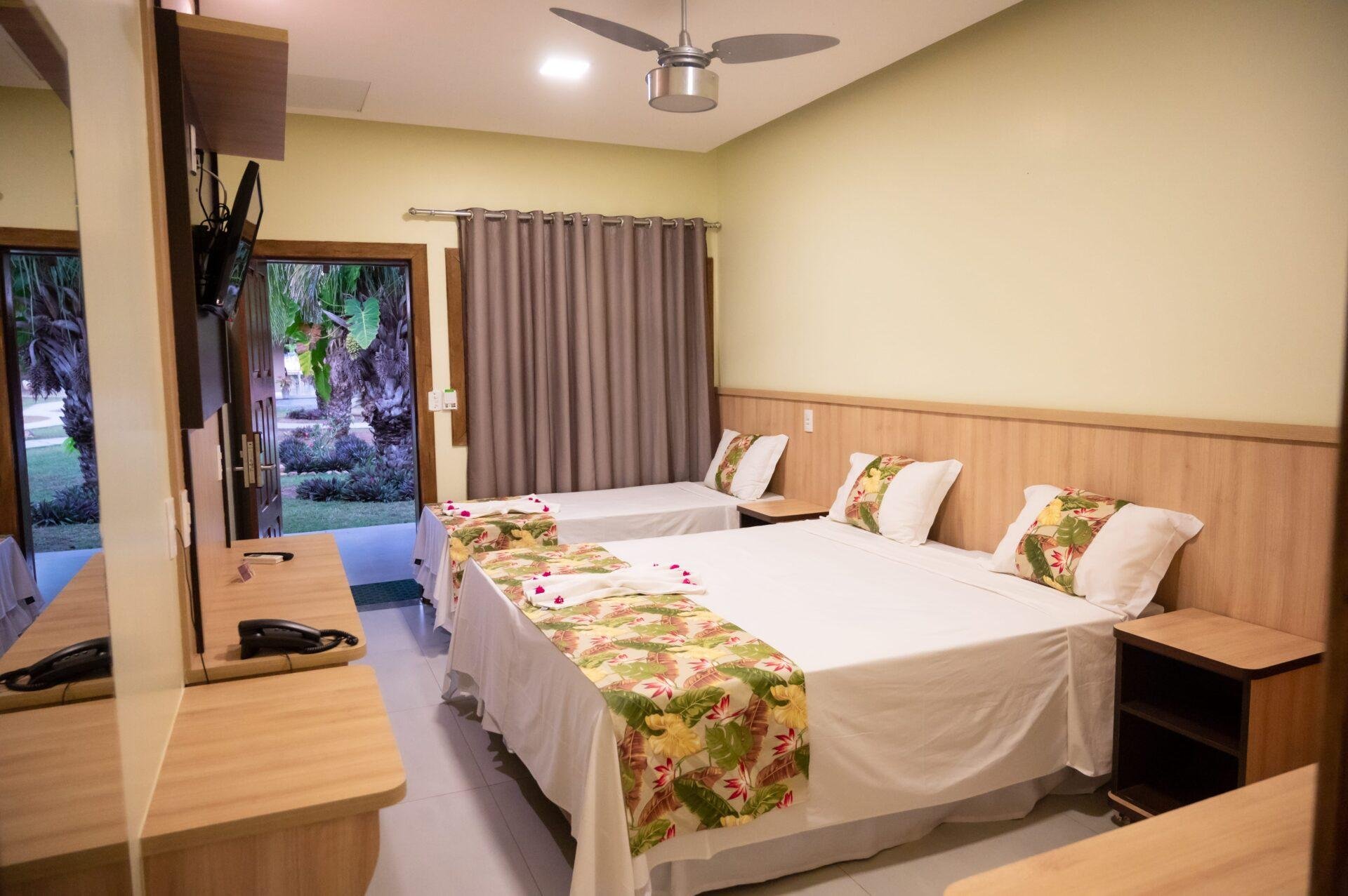 Tropical prints, fresh linens, and a garden just outside the door. Triple guest room on a pantanal jaguar budget safari, with two double beds dressed in tropical-print runners, ceiling fan, wall-mounted TV, and an open door revealing lush garden palms beyond