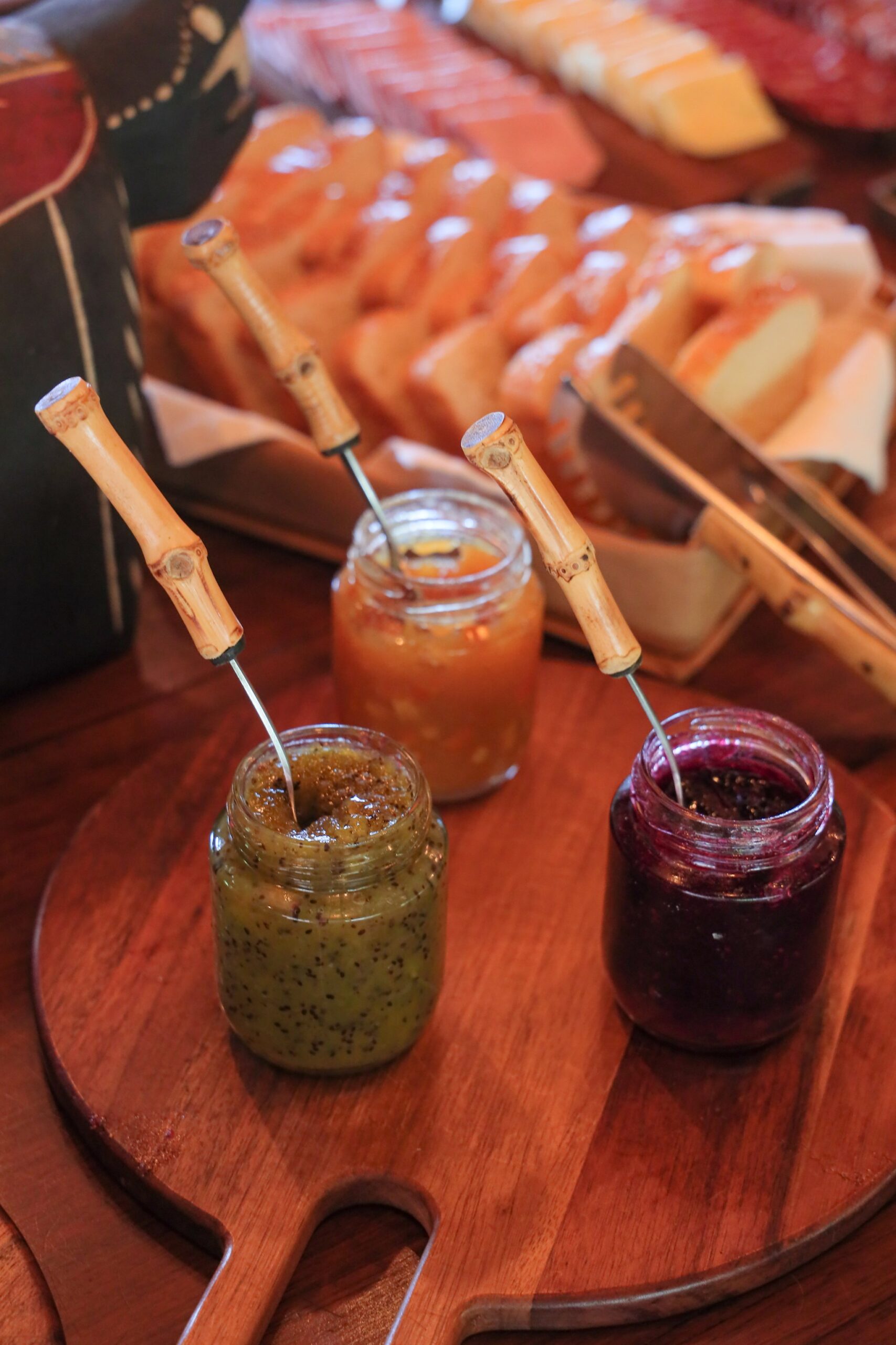 Three house-made preserves, a round board, and bread still warm from the kitchen. Photo: thiagolbrito Three glass jars of homemade fruit preserves — green, orange, and berry — with bamboo-handled spoons on a round wooden board, served alongside sliced bread and cheese at a pantanal luxury safari lodge