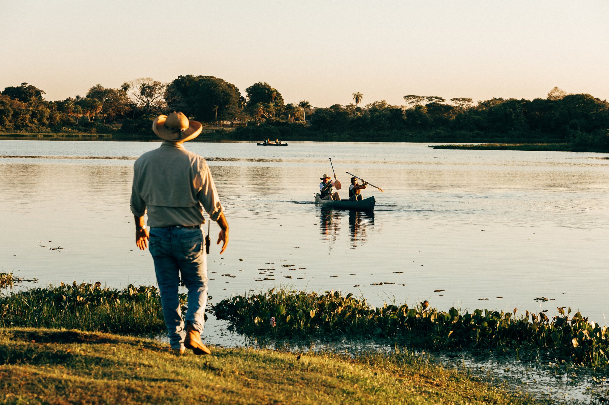Glassy water, a drifting canoe, and the last warmth of the day. A guide in a wide-brimmed hat watches from the bank as two guests paddle a canoe across a calm wetland lagoon during a pantanal luxury safari at golden hour