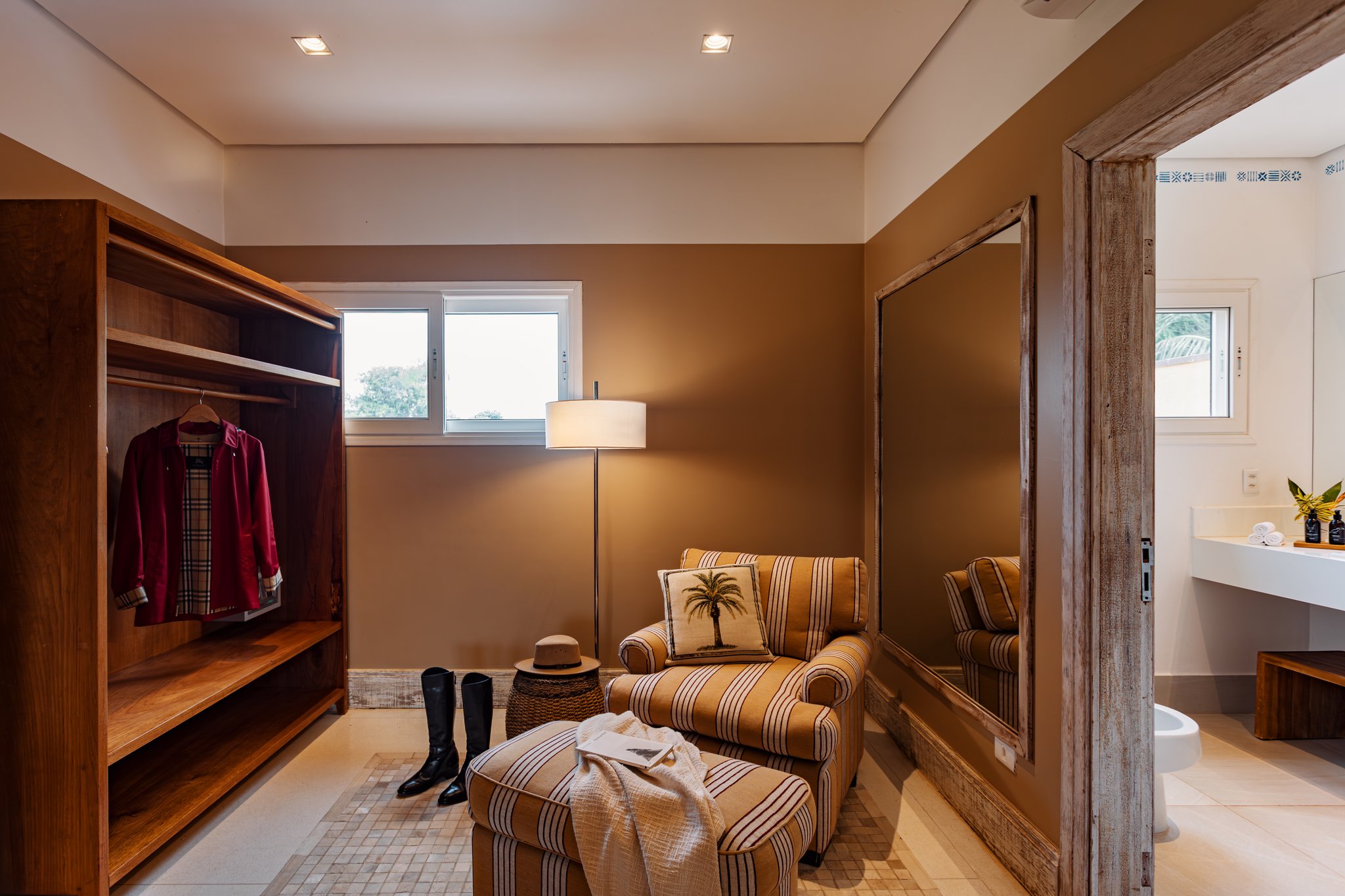 Every detail considered — from the open wardrobe to the palm-print cushion, your suite is ready before the safari begins. Photo: Felipe Castellari A warmly lit dressing area in a pantanal luxury safari lodge, featuring an open timber wardrobe, a striped armchair with a palm-motif cushion, riding boots, a straw hat, and an ensuite bathroom glimpsed through a rustic wooden doorframe