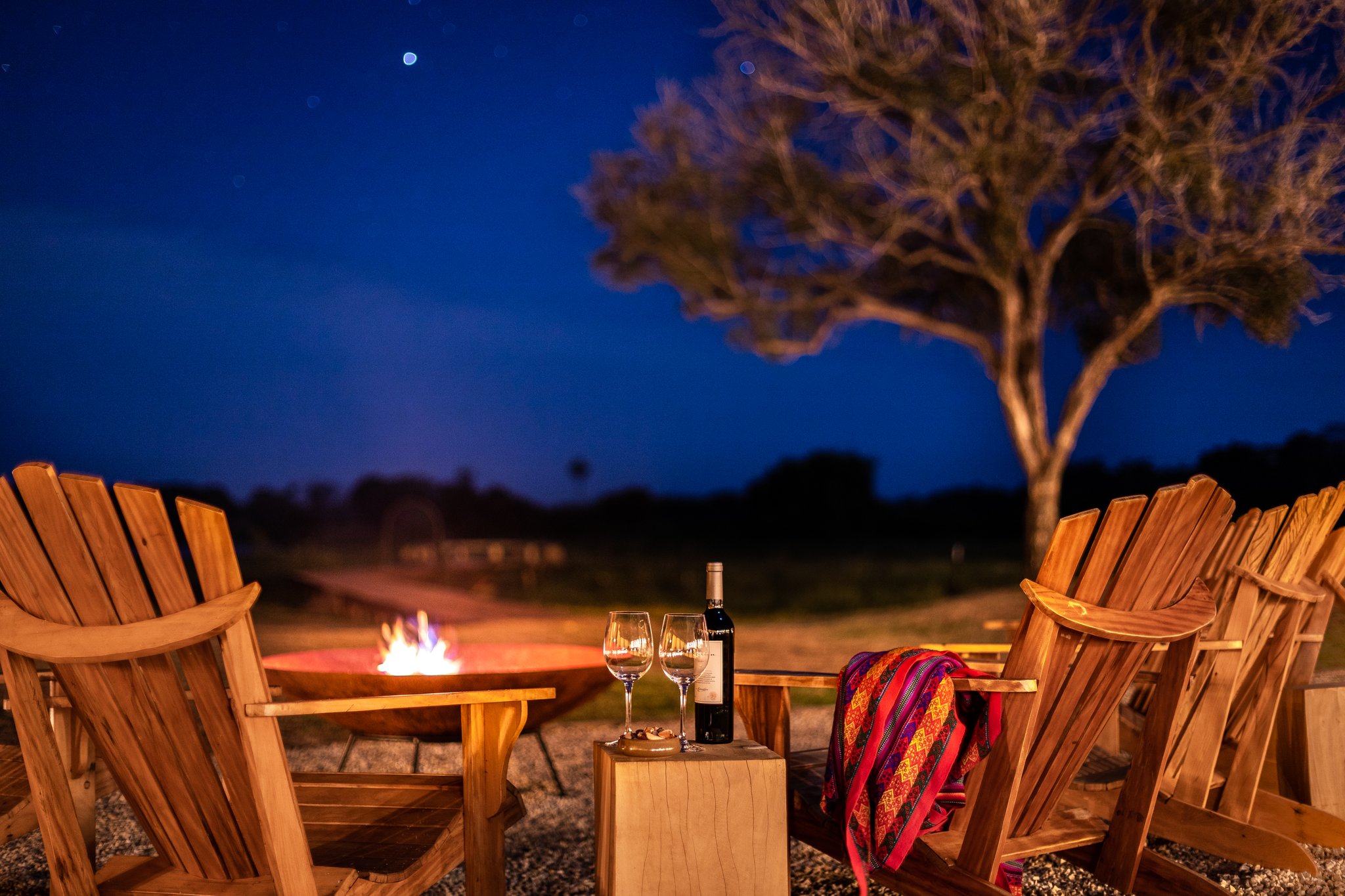 Wooden Adirondack chairs arranged around a glowing fire bowl at dusk during a pantanal luxury safari, with two wine glasses and a bottle on a log side table under a deep blue sky