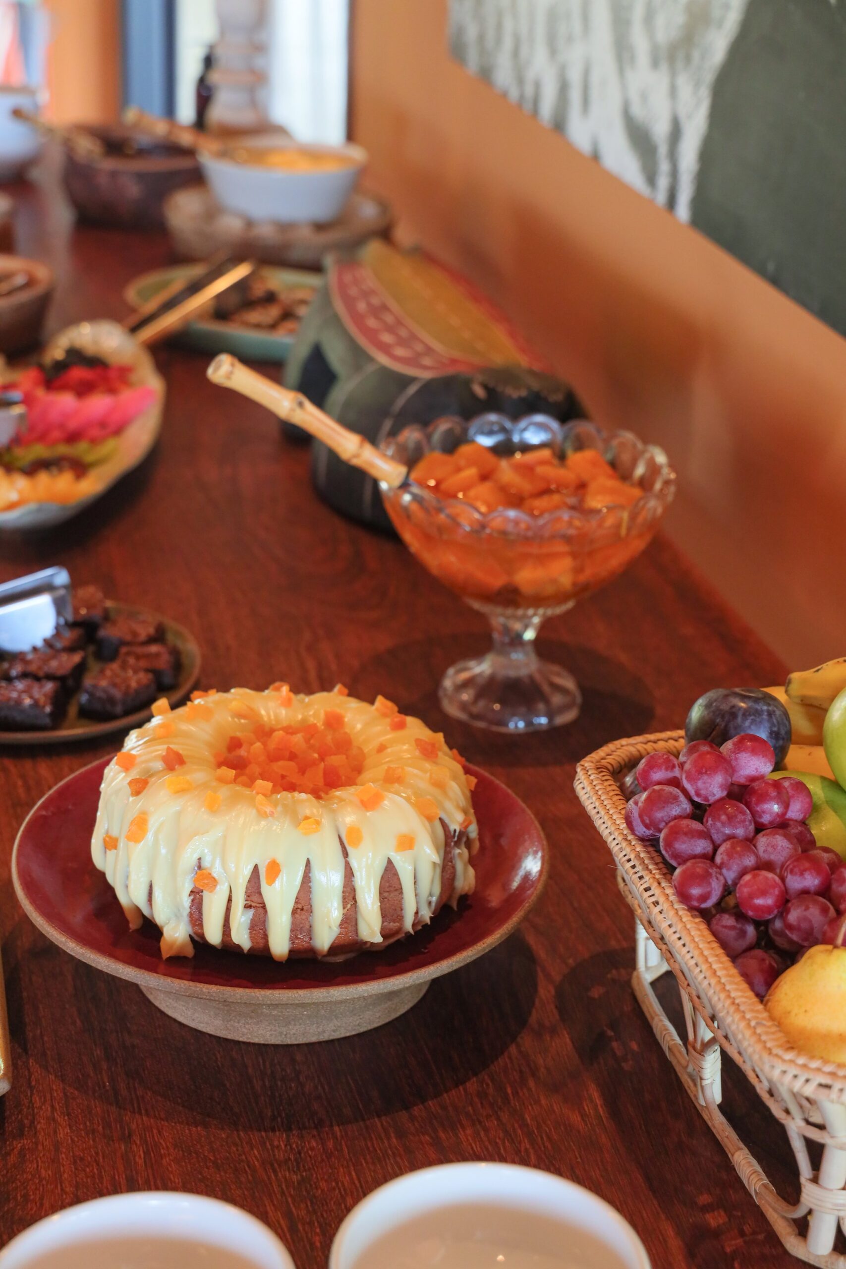 Homemade, unhurried, generous — the lodge table before the day begins. Photo: thiagolbrito A glazed bundt cake topped with diced papaya sits on a red ceramic stand at a pantanal luxury safari lodge breakfast buffet, surrounded by fresh grapes, diced fruit, and brownies