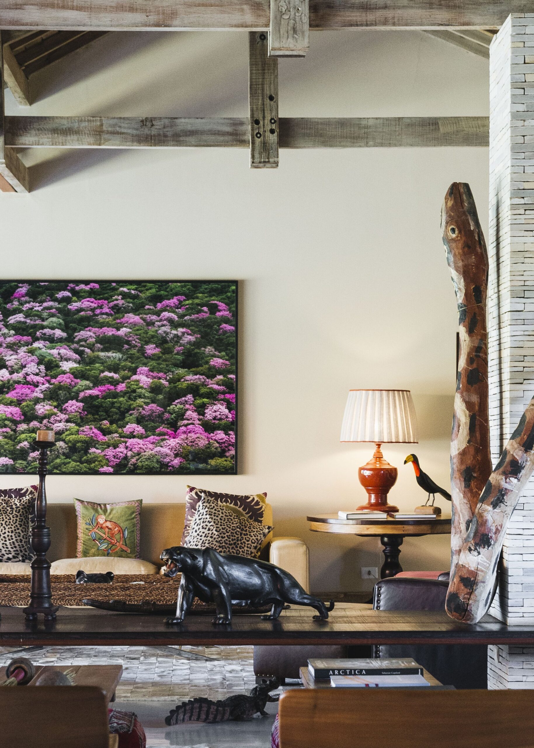 Rustic-chic lodge interior with exposed timber beams, a black panther sculpture on a console table, wildlife-print cushions, a toucan figurine beside a warm lamp, and a large nature photograph of pink-blooming trees on the wall — the refined character of a pantanal luxury safari lodge