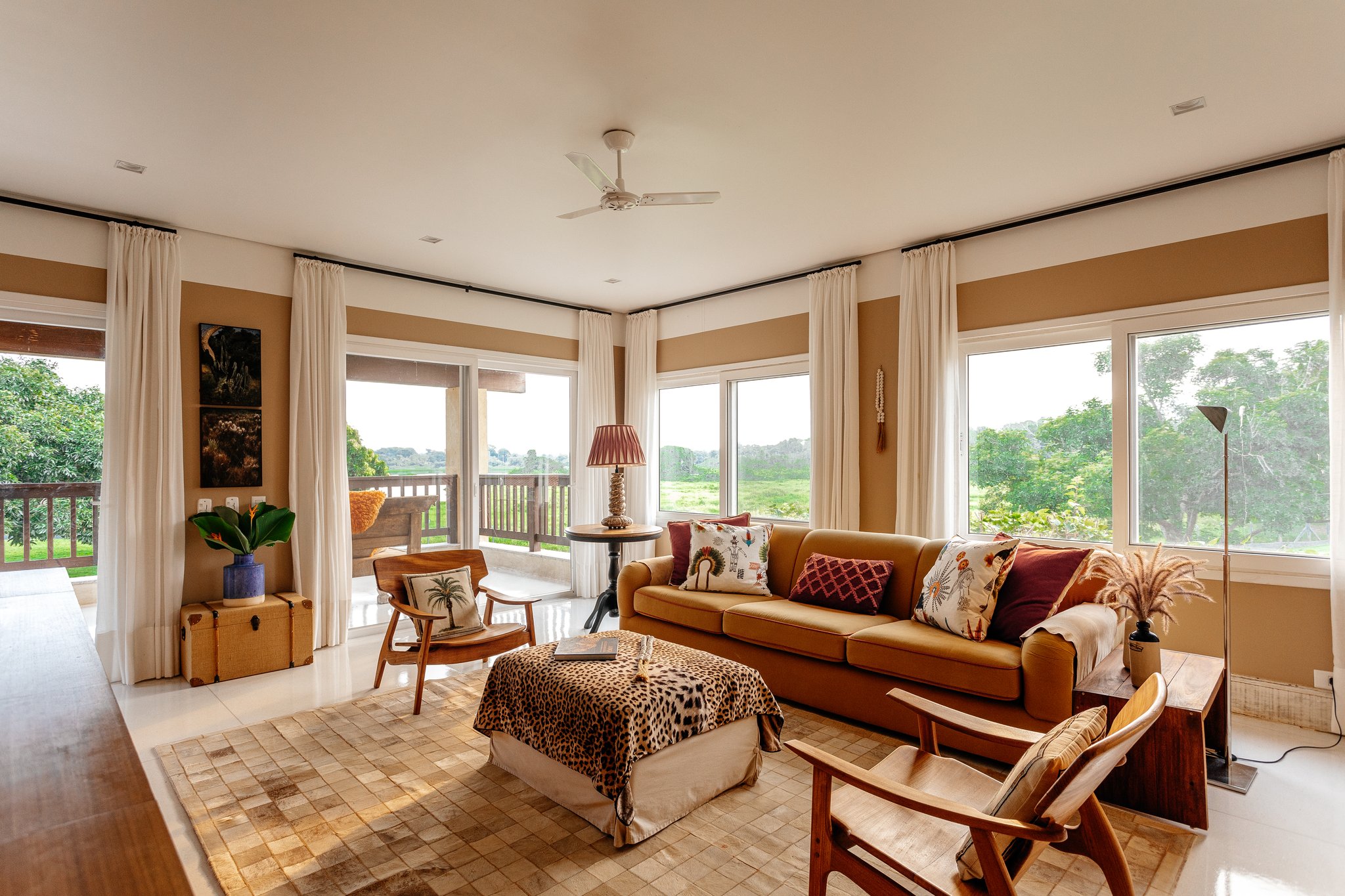 Warm earth tones and sweeping wetland views — comfort that matches the wildness outside. Photo: Felipe Castellari Sunlit living room at a pantanal luxury safari lodge, featuring a caramel sofa with patterned cushions, a leopard-print ottoman, timber armchairs, and panoramic windows overlooking open wetland vegetation