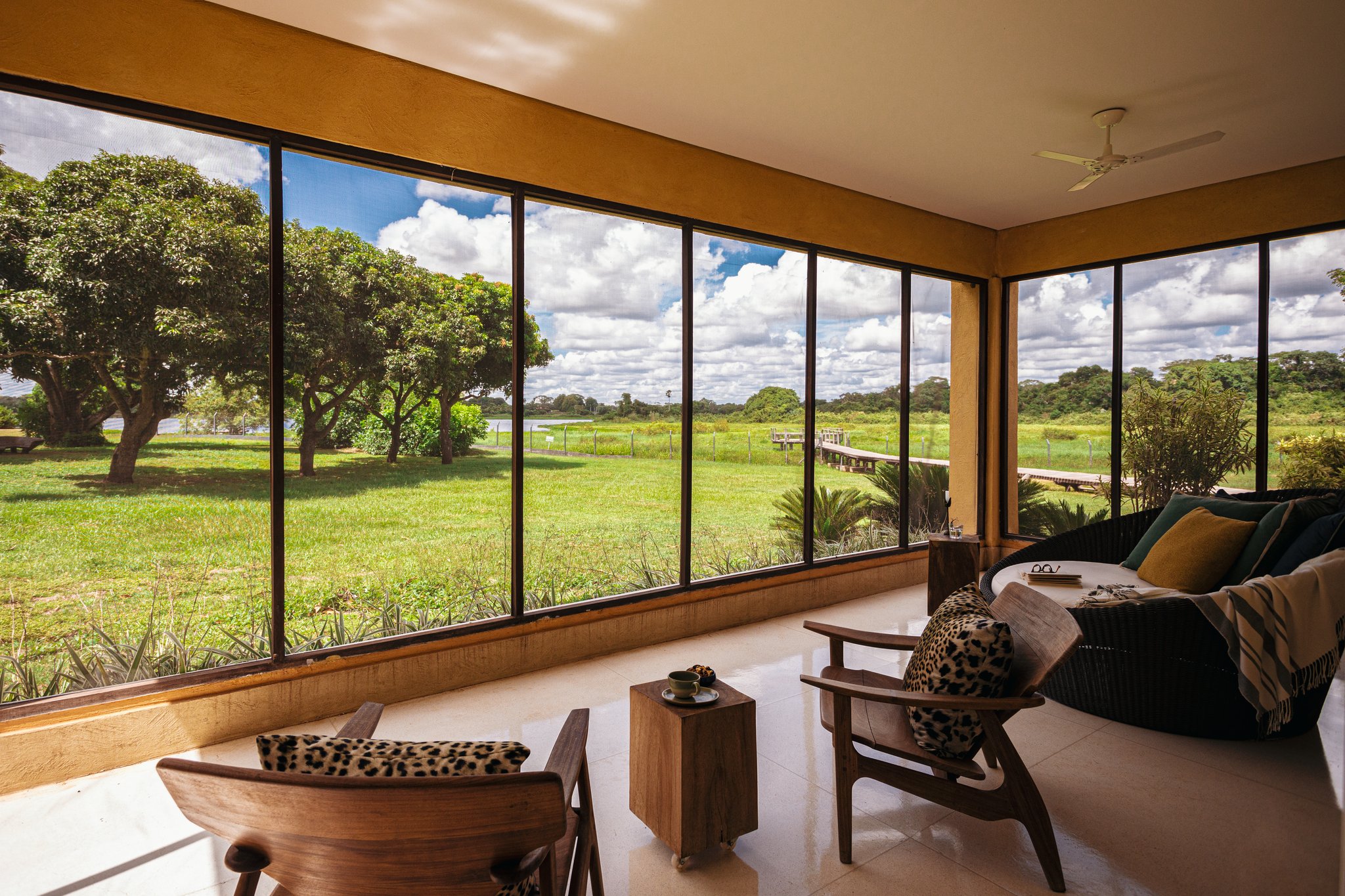 Leopard-print cushions, warm timber, and a vast wetland stretching to the horizon — comfort and wilderness in one frame. Photo: Felipe Castellari Elegant lounge area with floor-to-ceiling windows overlooking a green wetland meadow and wooden boardwalk at a pantanal luxury safari lodge